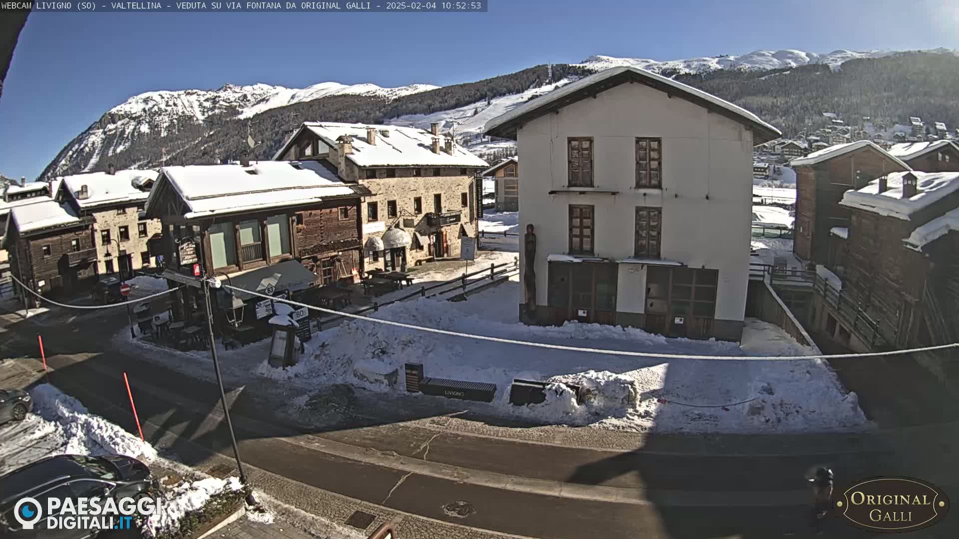 A snow-covered village square with several buildings, some cars, and a sunny, clear sky with mountains in the background.