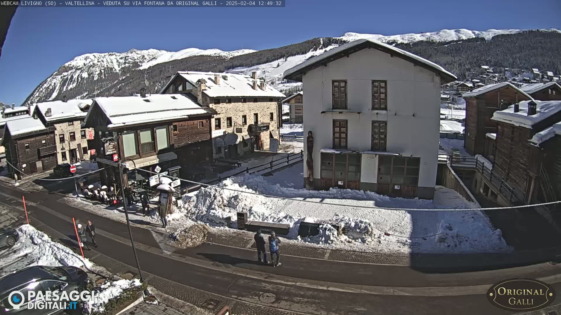 A sunny, snow-covered village square with several buildings, some people walking, and cars parked along a street.