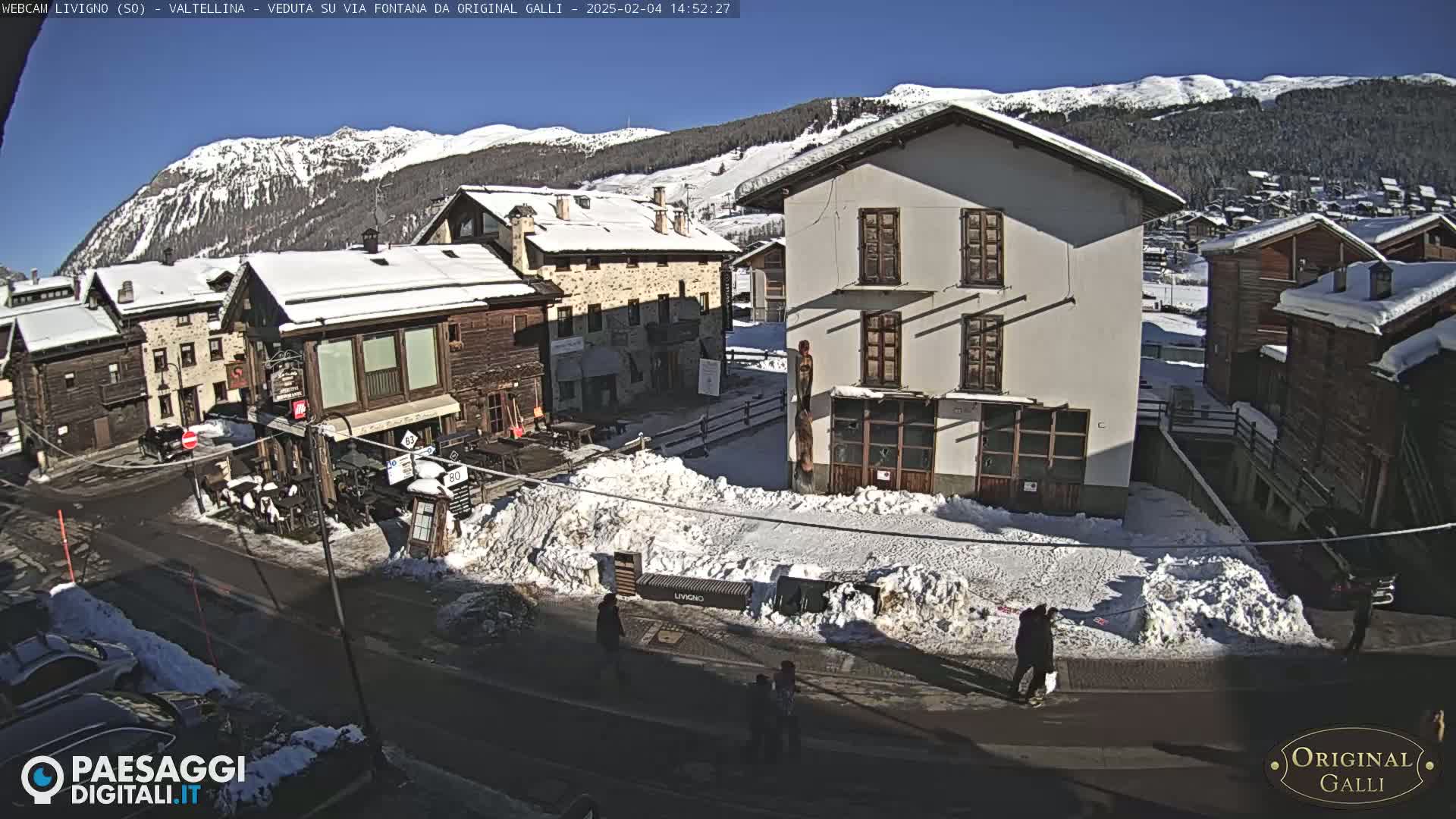 A snow-covered village street scene under a bright, sunny sky, with mountains in the background.