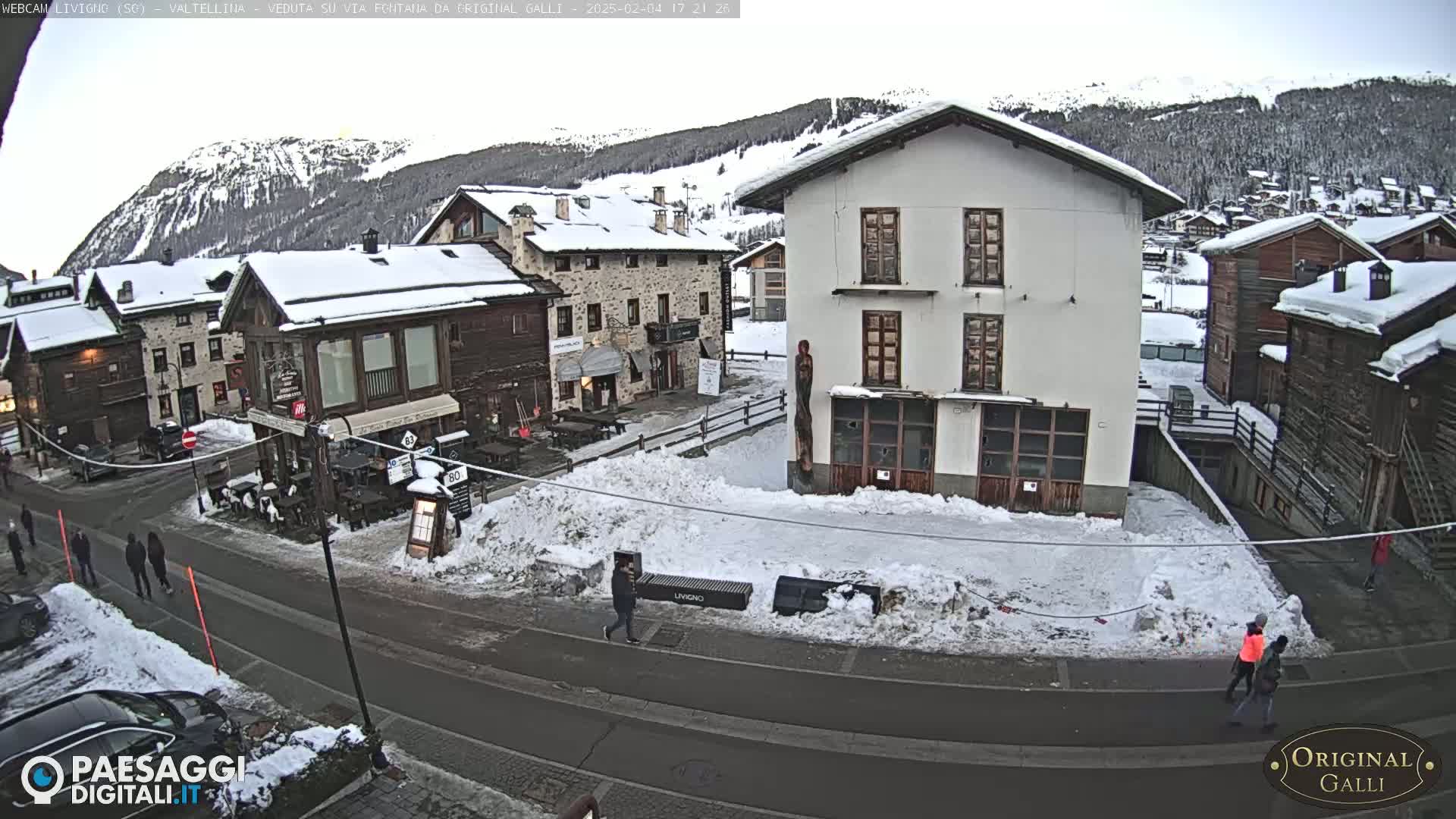 A snow-covered village street scene with several buildings, some pedestrians, and a mountainous background under a cloudy sky.
