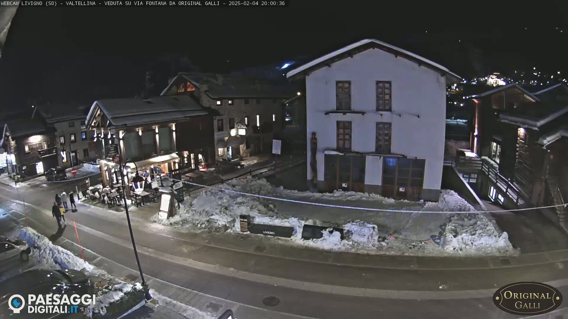 A nighttime street scene in a snow-covered village shows several buildings, a few people walking, and cars parked along the road.