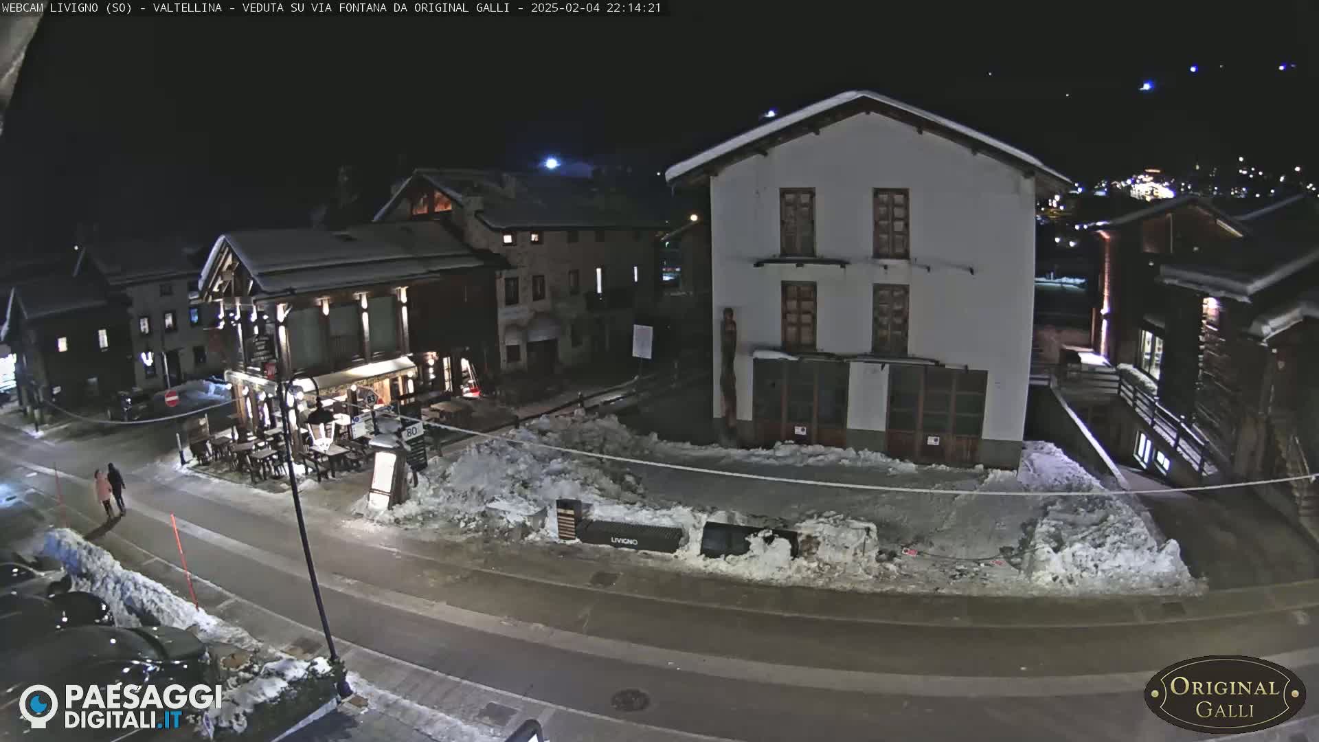 A snow-covered street scene at night shows several buildings, one of which is a restaurant with outdoor seating, and a couple walking down the street.