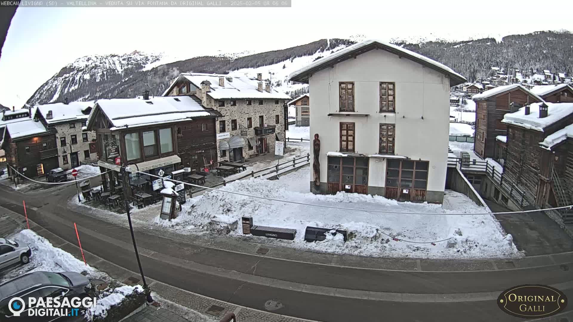 A snow-covered village square with several buildings, some with snow on their roofs, is viewed from above on a cloudy day with snow-covered mountains in the background.