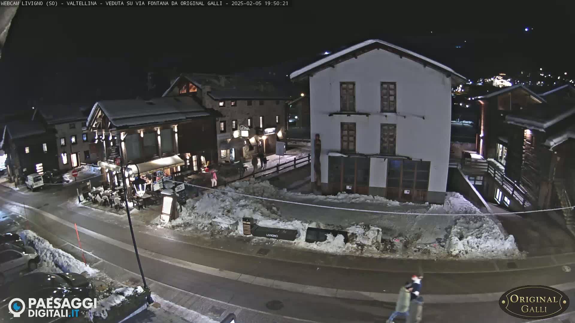 A snow-covered street scene at night shows several buildings, a few people walking, and some parked cars.