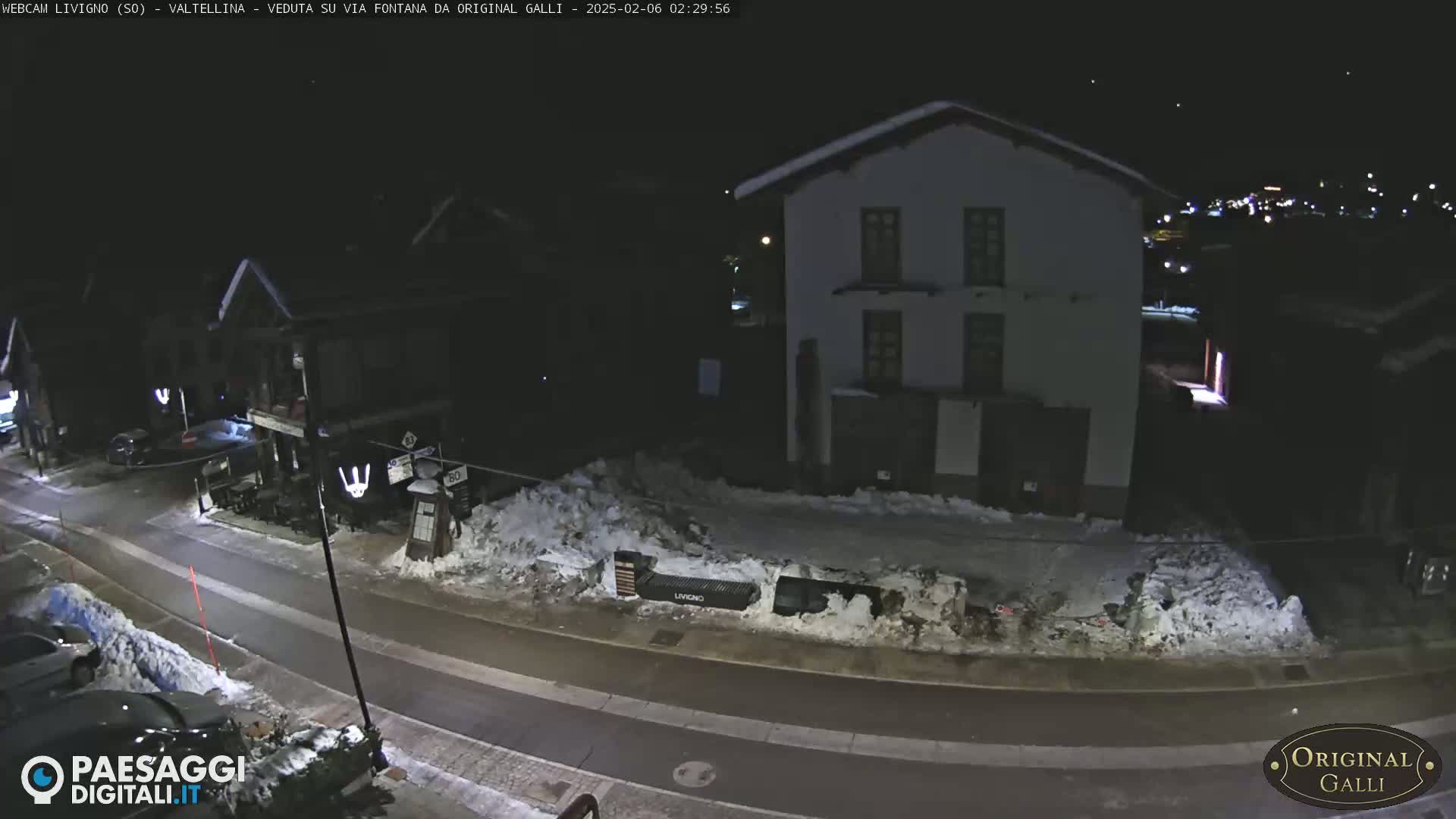 A snow-covered street at night, lit by streetlights, shows a two-story building and parked cars.