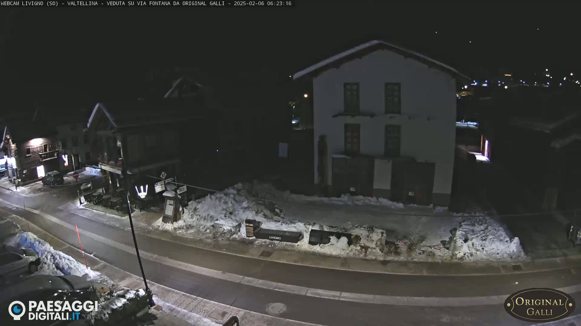 A nighttime street scene shows snow-covered ground and buildings, with a few cars parked along the road.