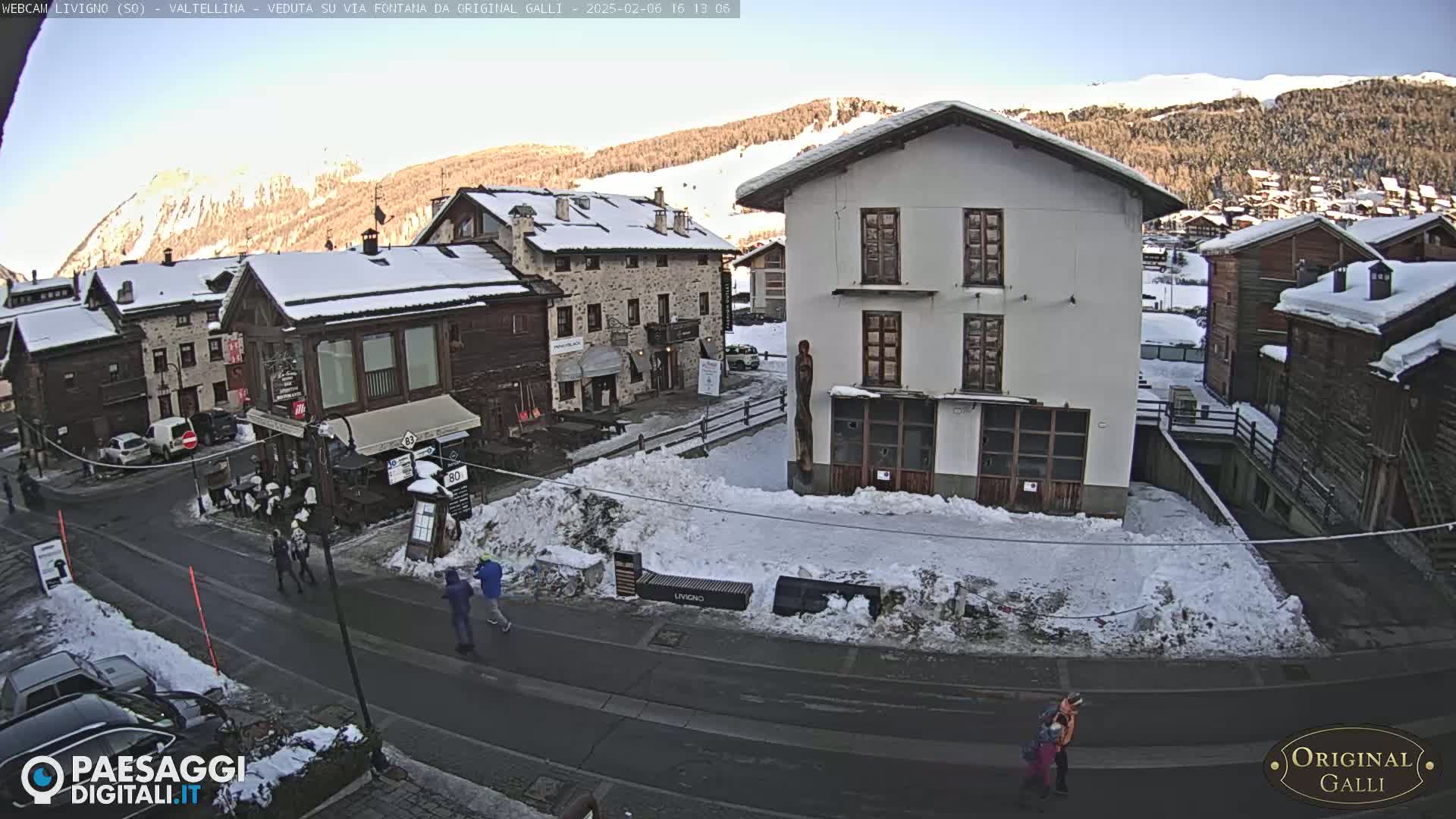 A snow-covered village street scene shows several buildings, some pedestrians, and cars parked along the road, with sunny, clear skies and snow-capped mountains in the background.