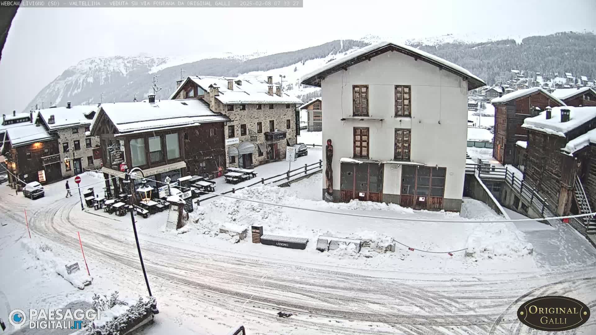 A snow-covered village square with several buildings, some with outdoor seating areas, is visible under a lightly snowing sky, with mountains in the background.
