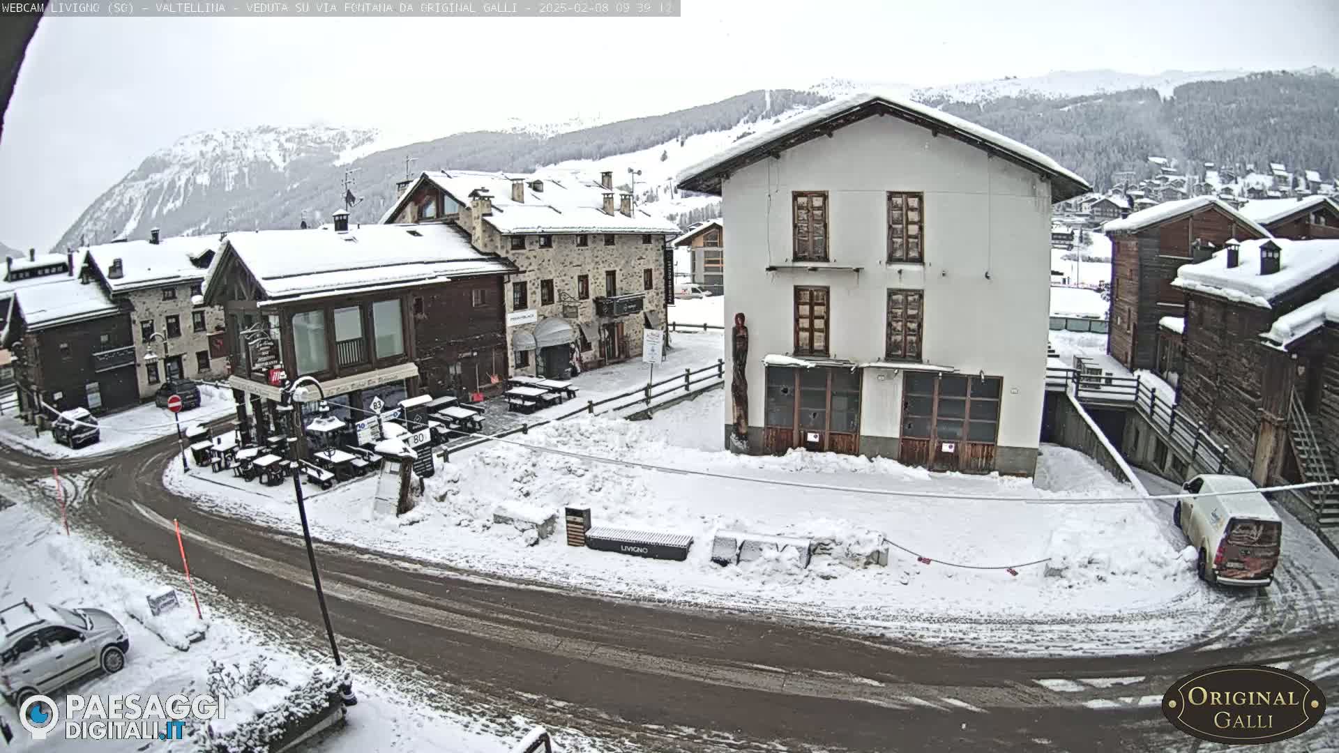 A snow-covered village square with several buildings, a few parked cars, and outdoor seating areas in a mountainous region under an overcast sky.