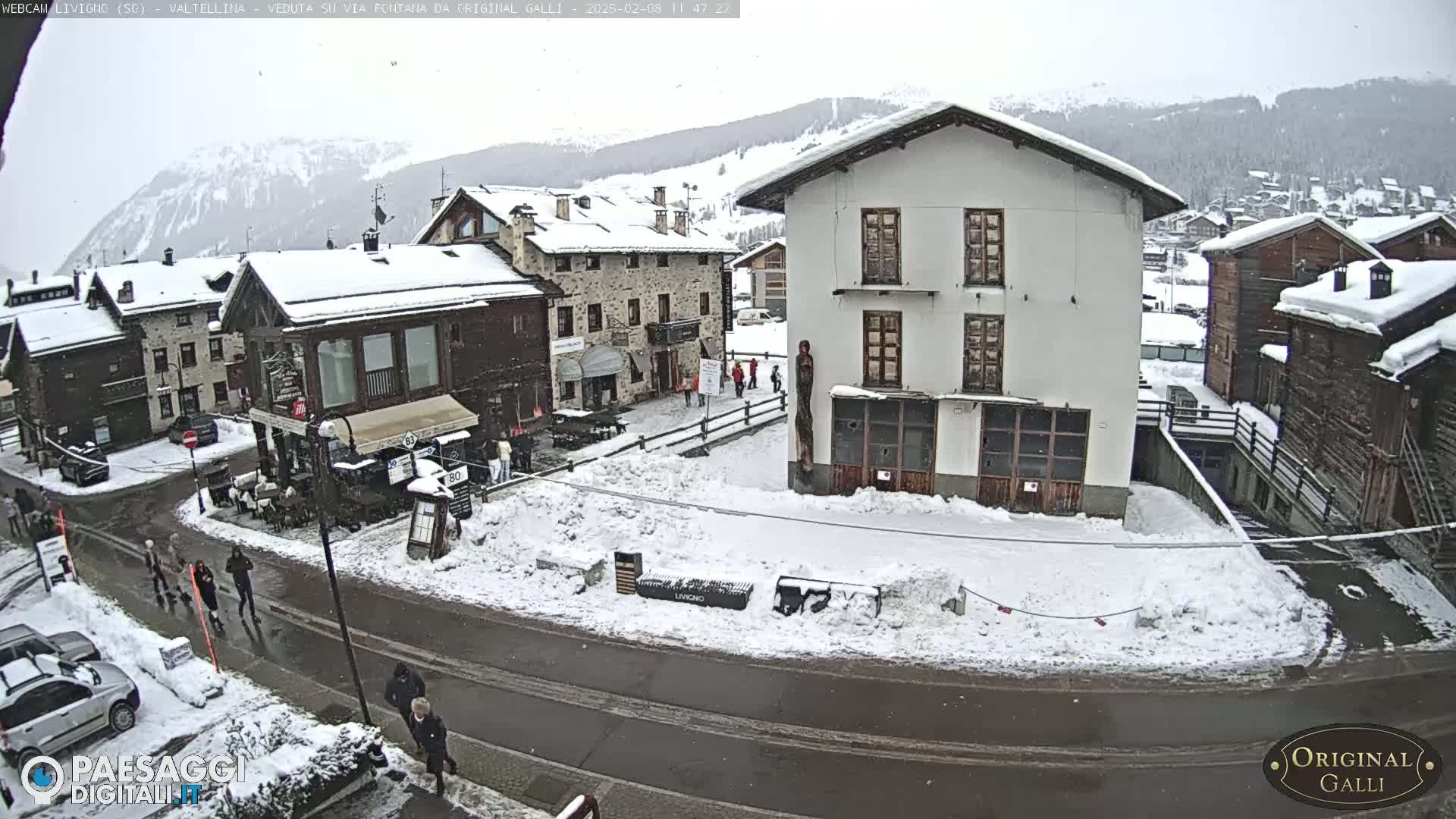A snow-covered village square with several buildings, a road, and a few pedestrians, under a lightly snowing sky.