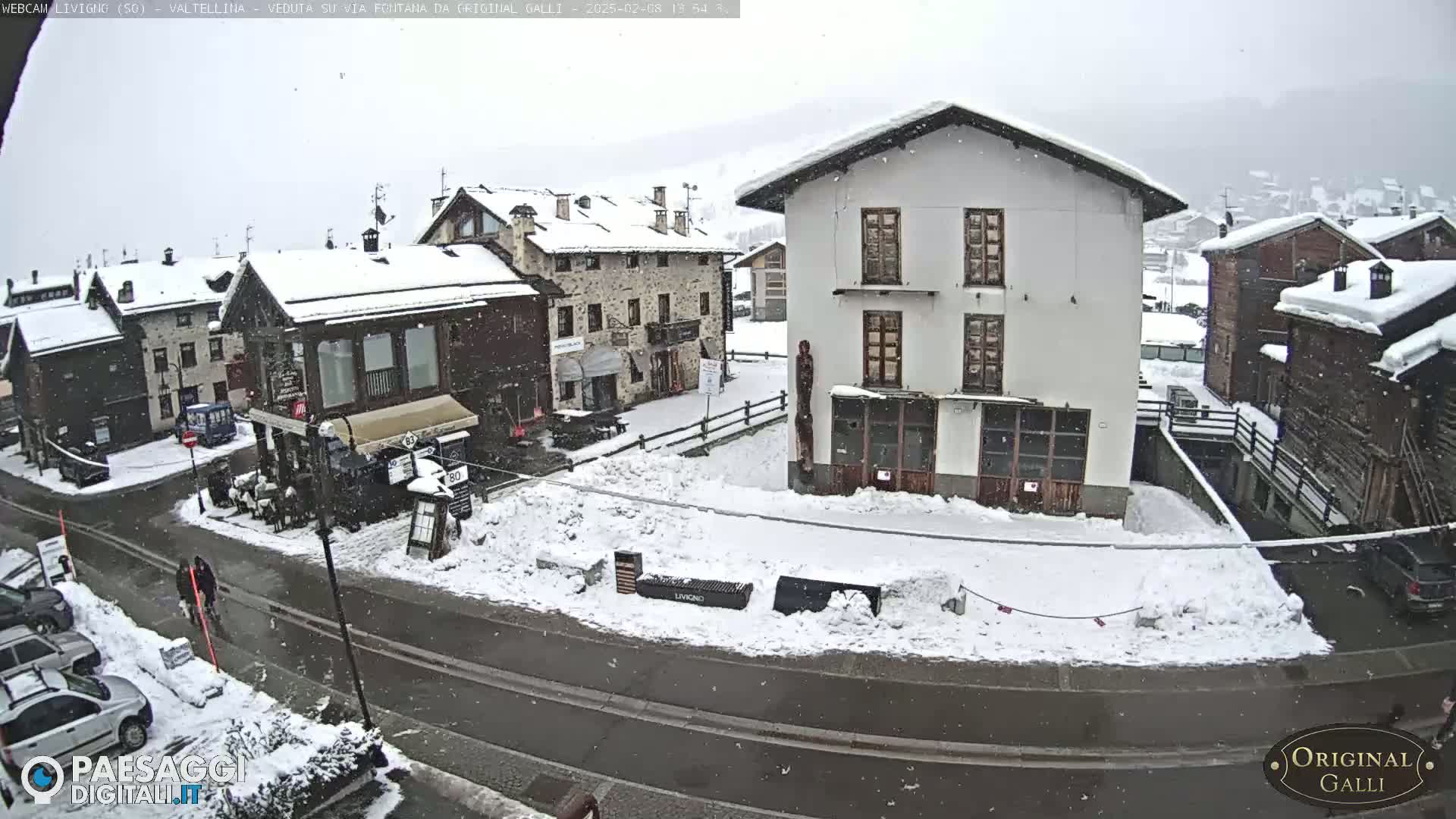 A snow-covered town square with several buildings, a street, and some parked cars under a light snowfall.