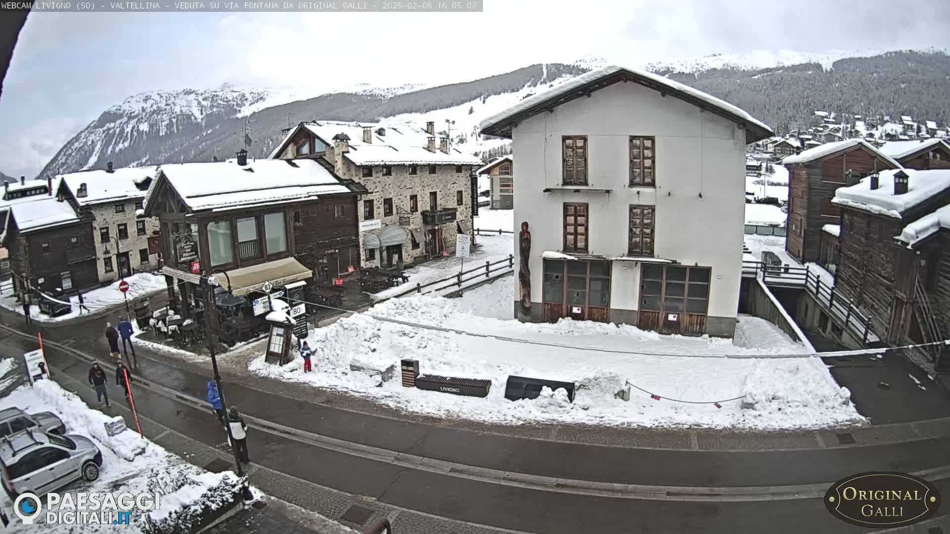 A snow-covered village street scene shows several buildings, some people walking, and parked cars under an overcast sky.