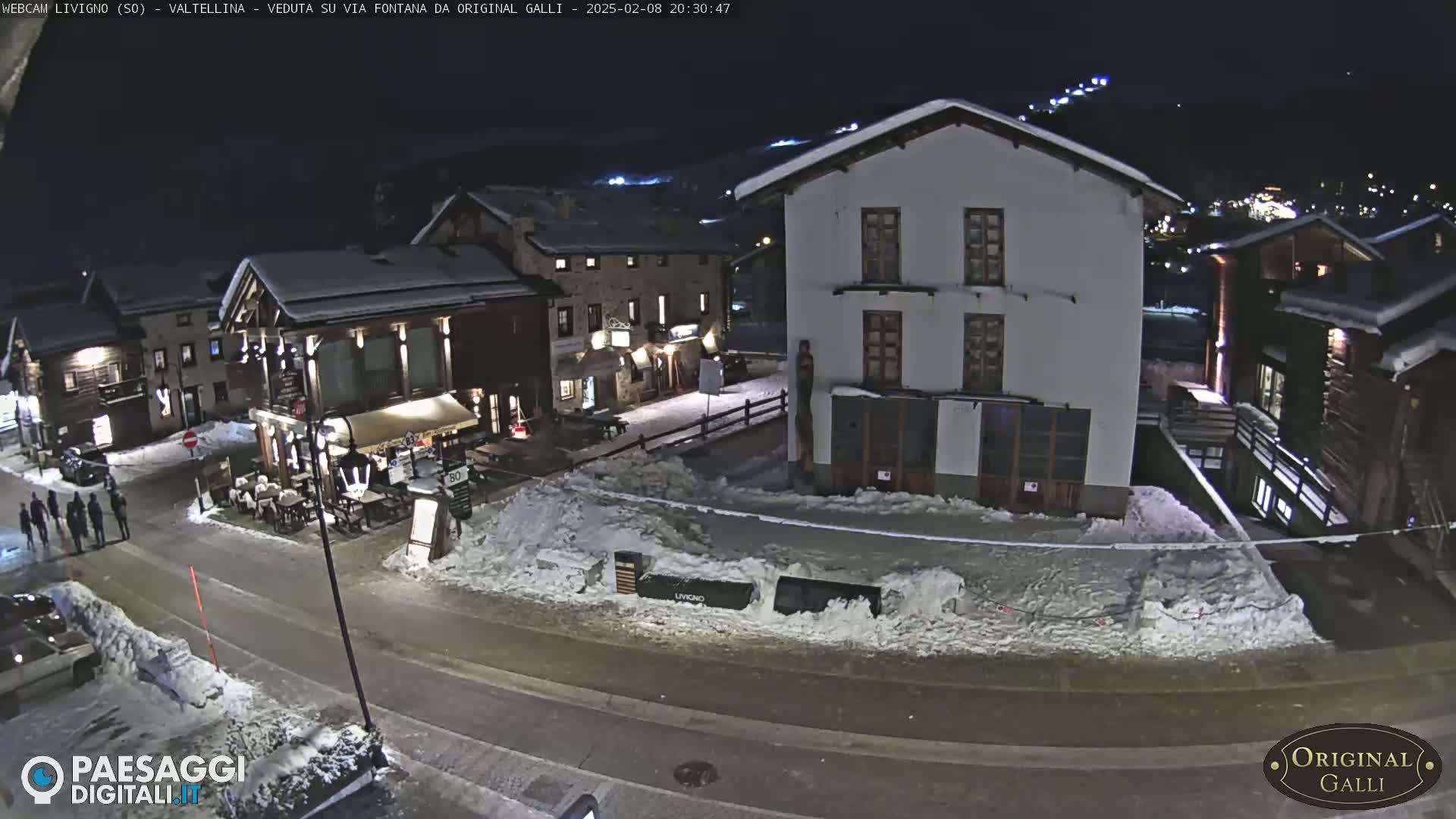 A snow-covered village square at night, illuminated by streetlights, with several buildings and a few people visible.