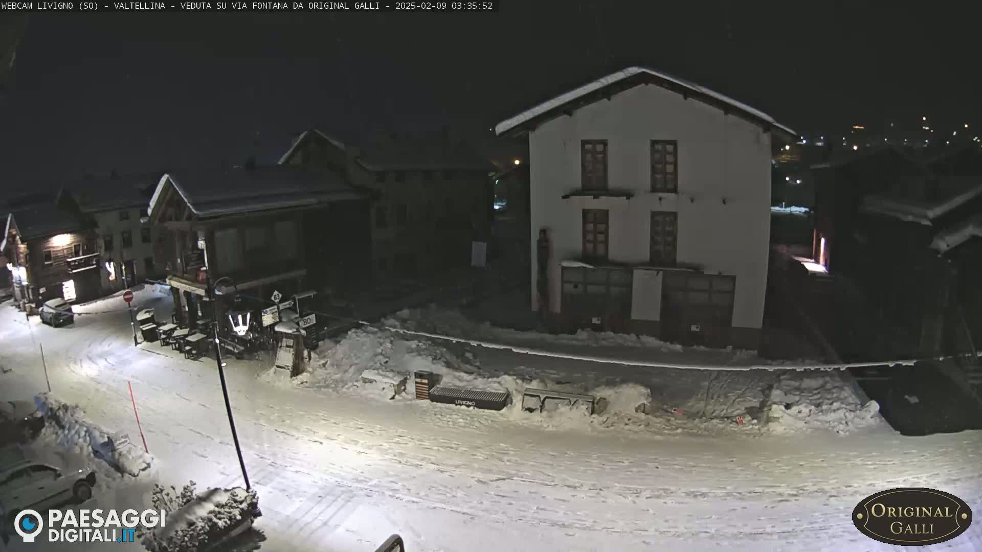 A snow-covered street at night, illuminated by streetlights, with several buildings visible in a small village.