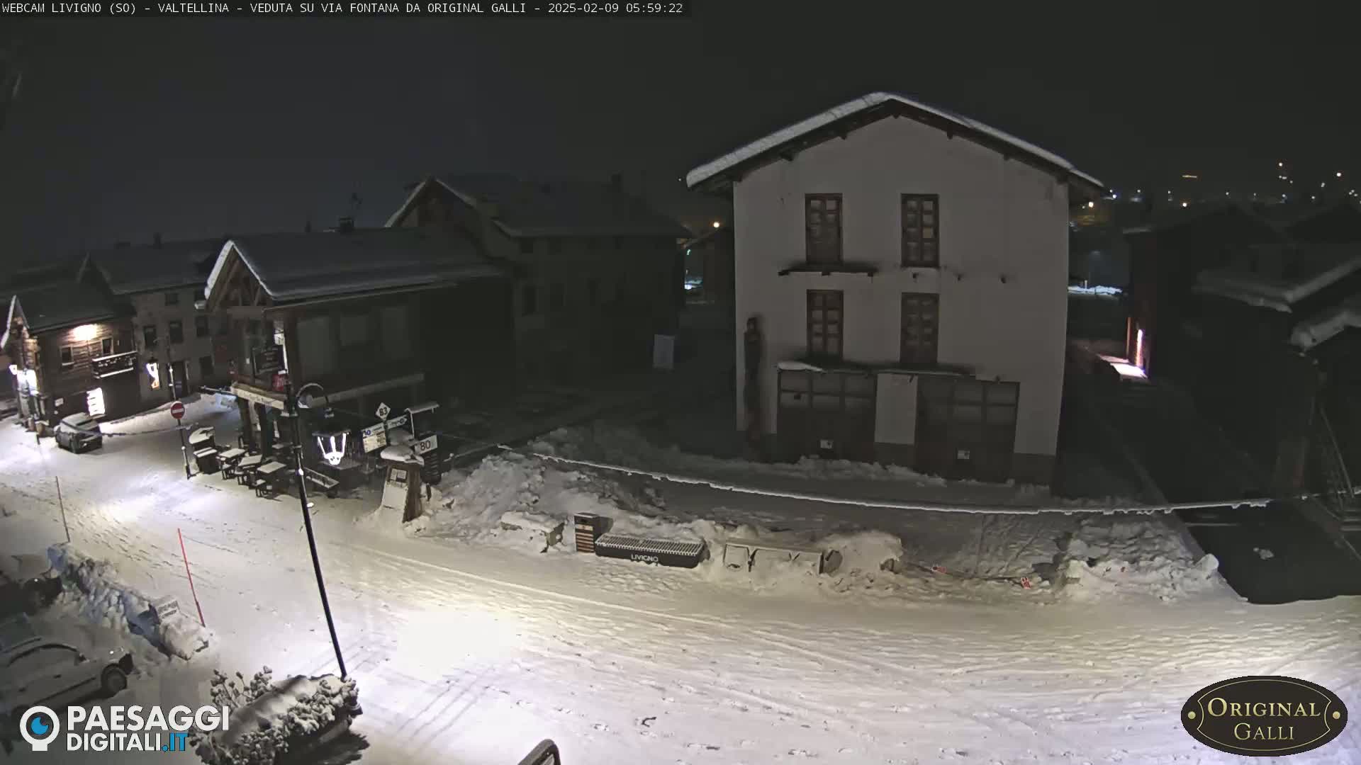A snow-covered street scene at night, showing several buildings illuminated, with some parked cars and a few outdoor tables visible.