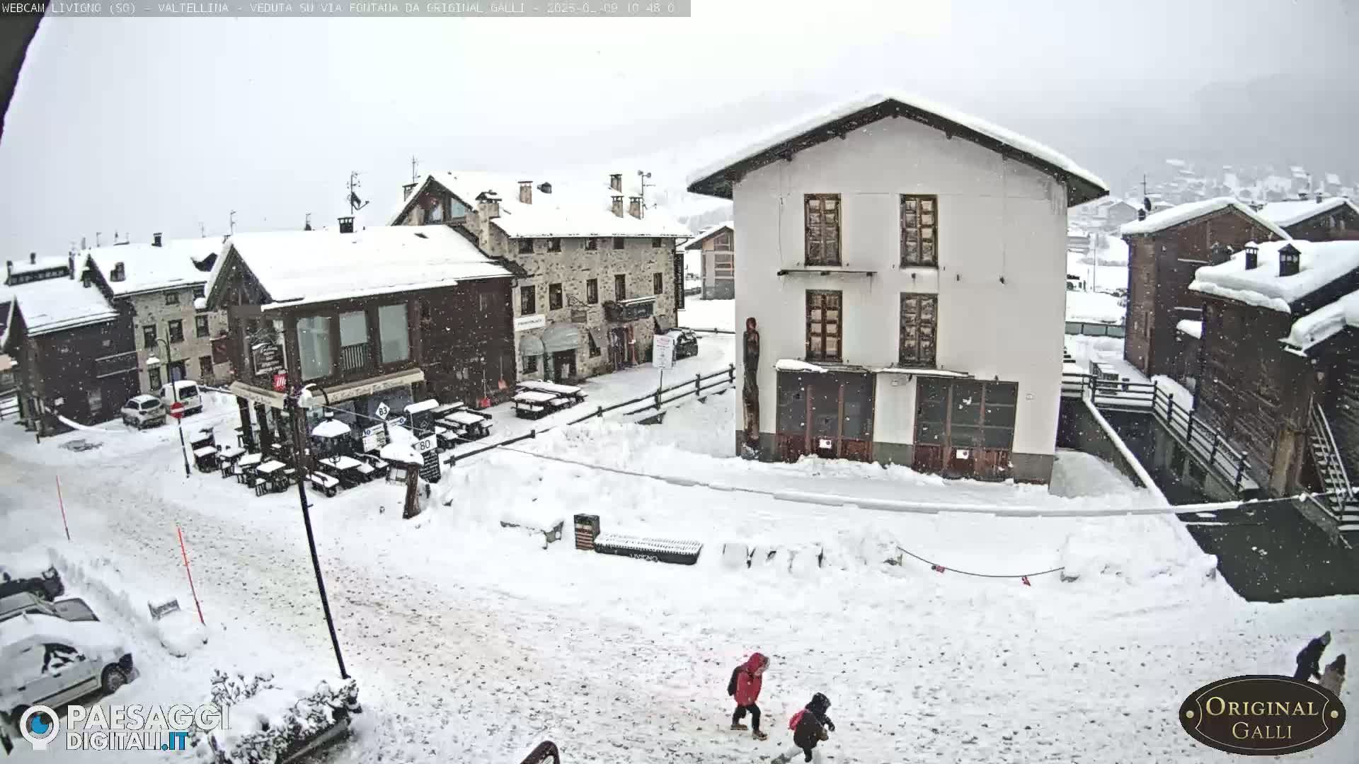 A snow-covered village square with several buildings, some cars parked in the snow, and two children walking in the falling snow.