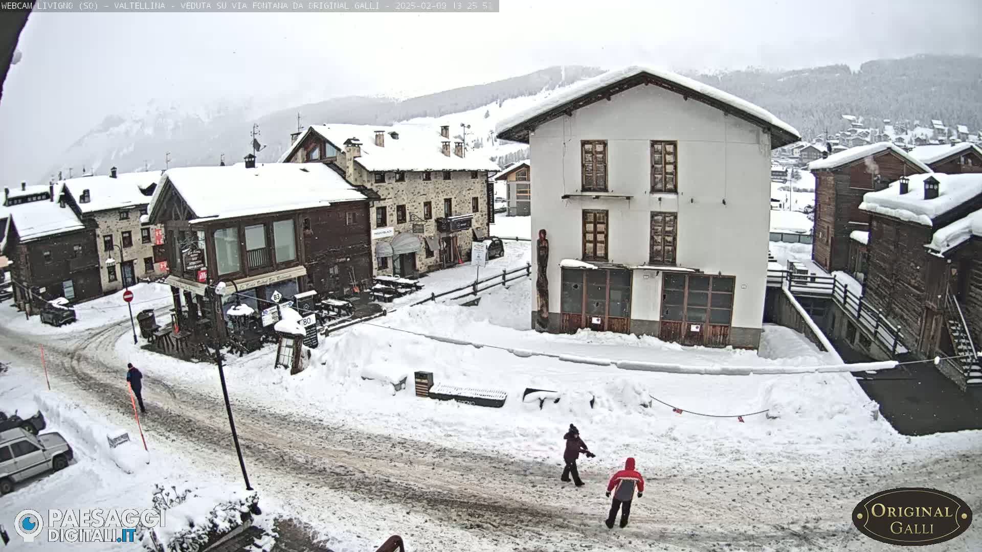A snow-covered village street scene, with several buildings and a few people walking, under an overcast sky.
