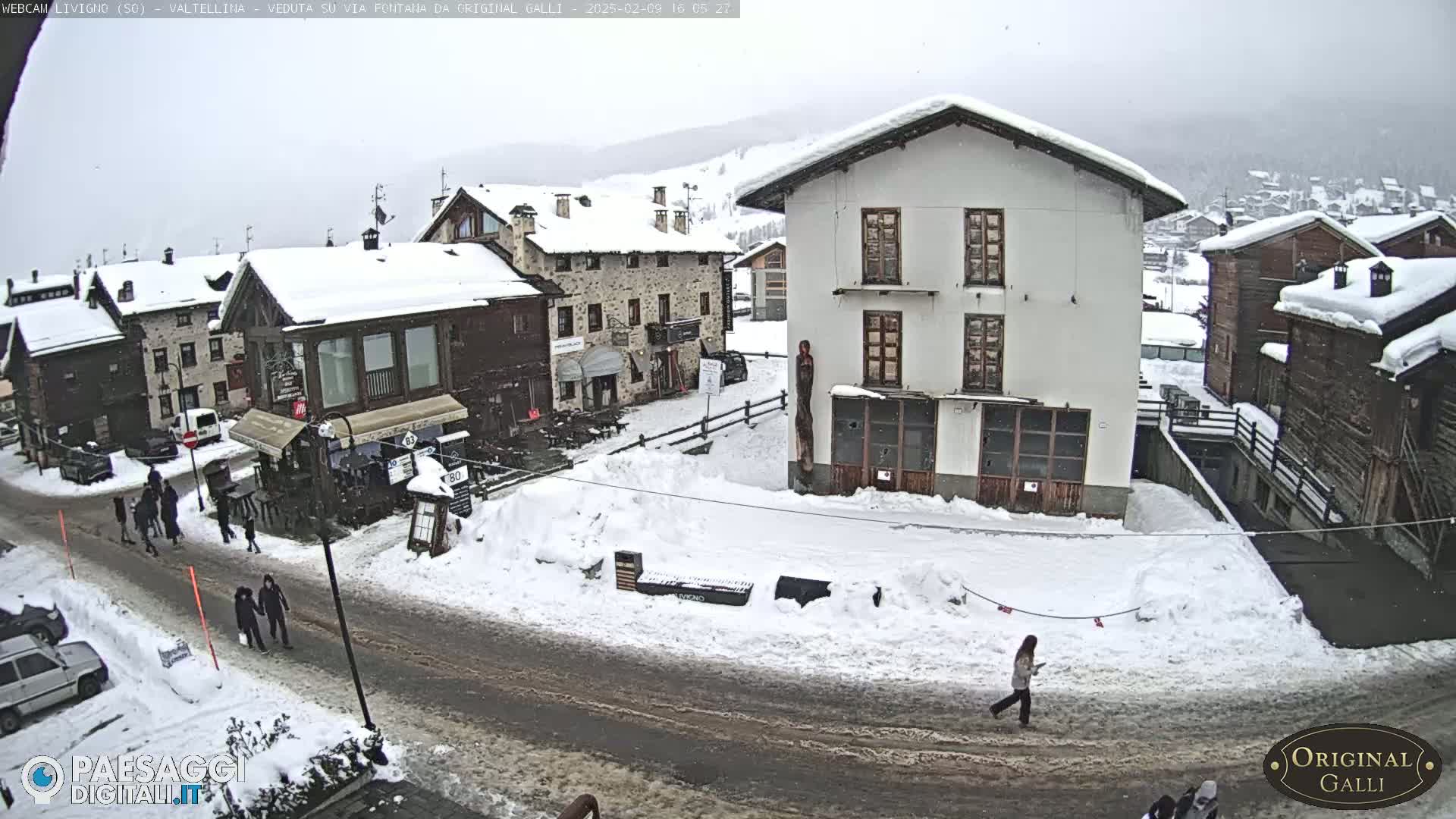 A snow-covered village street scene with several people walking, cars parked, and buildings covered in snow under an overcast sky.