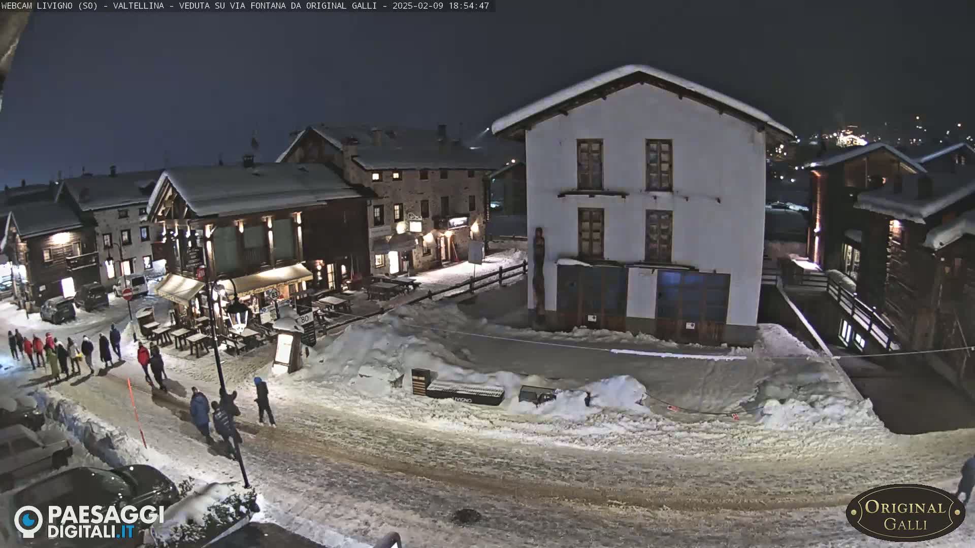A snow-covered village street at night, with several people walking and buildings illuminated.