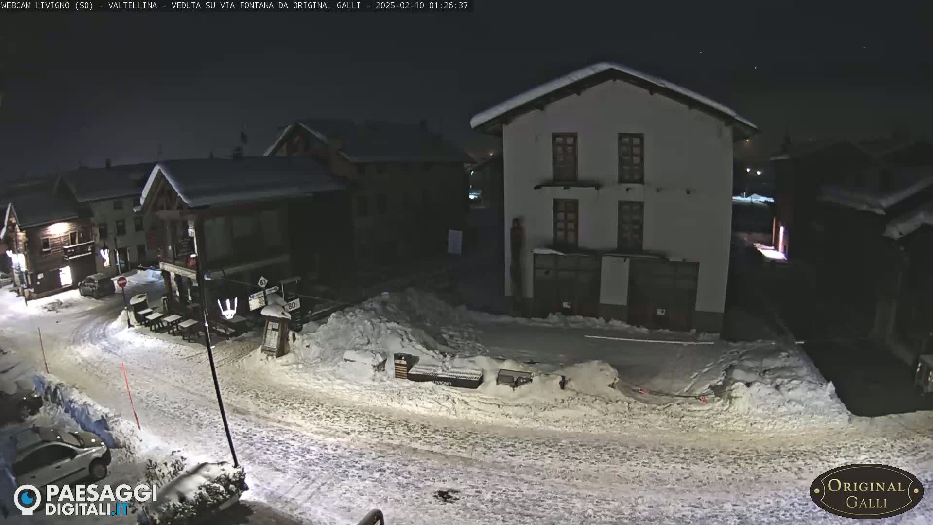 A snow-covered village street at night is illuminated by lights from buildings, with cars parked along the side.