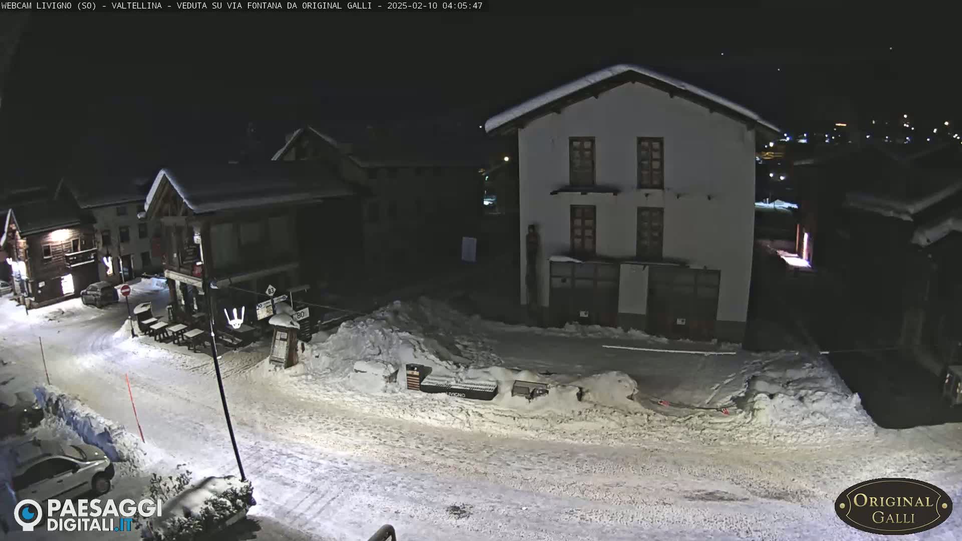 A snow-covered street at night in a village, with several buildings illuminated, shows cars parked along the sides of the road.