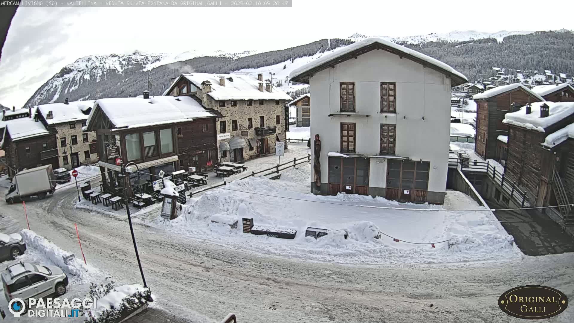 A snow-covered village square with several buildings, some cars, and outdoor seating areas under an overcast sky.