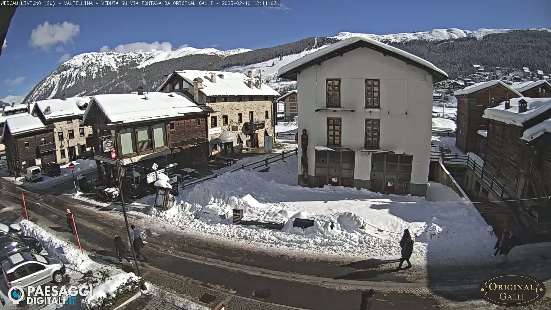 A snow-covered village street scene in the mountains on a sunny day, with several buildings and a few pedestrians.