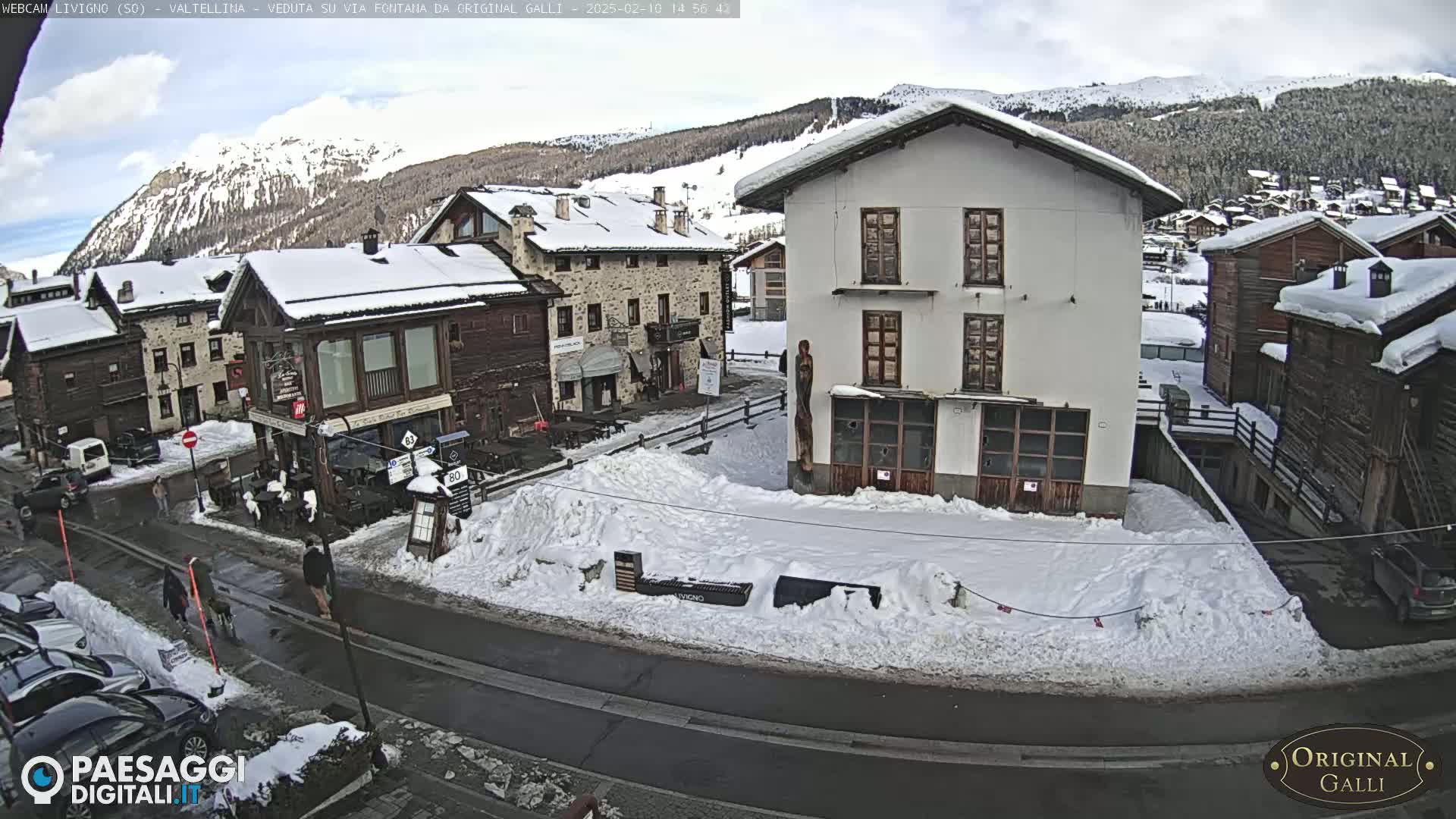 A snow-covered village scene shows several buildings, a street with pedestrians, and parked cars, all under a partly cloudy sky.