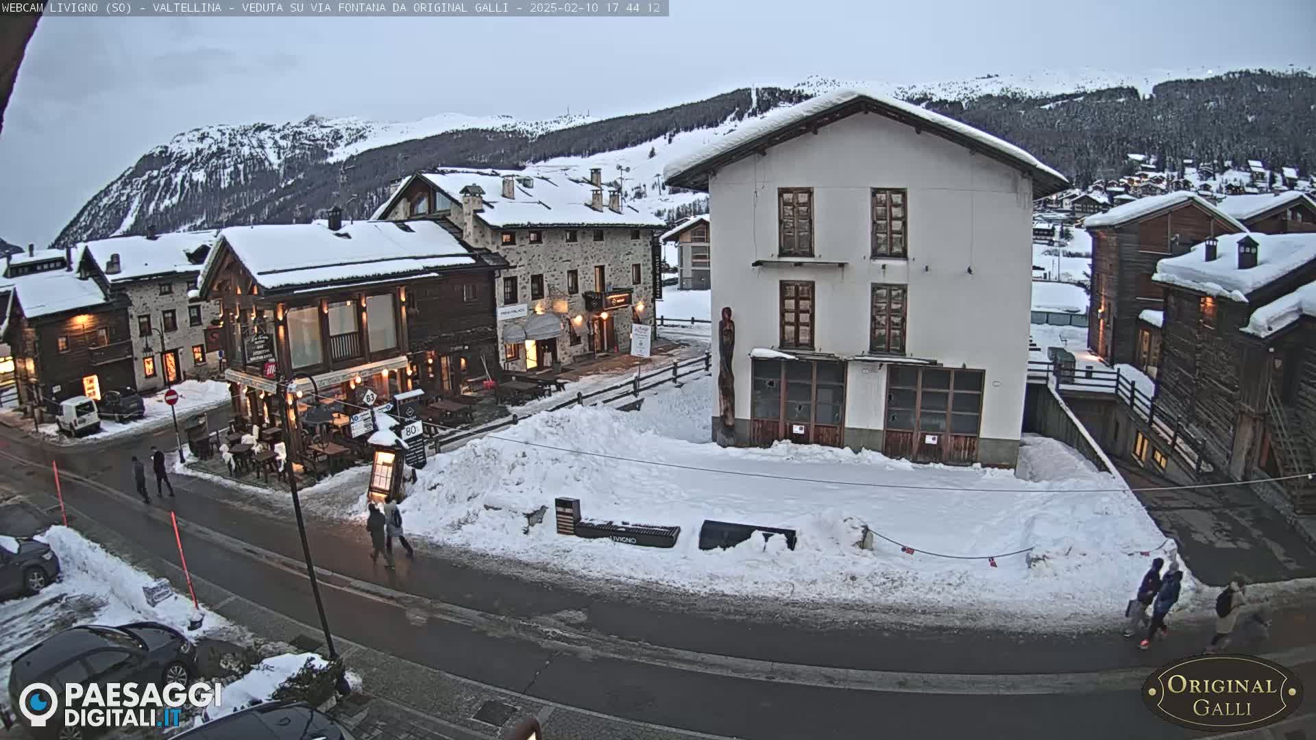 A snow-covered village street scene at dusk, with several buildings and a few pedestrians.