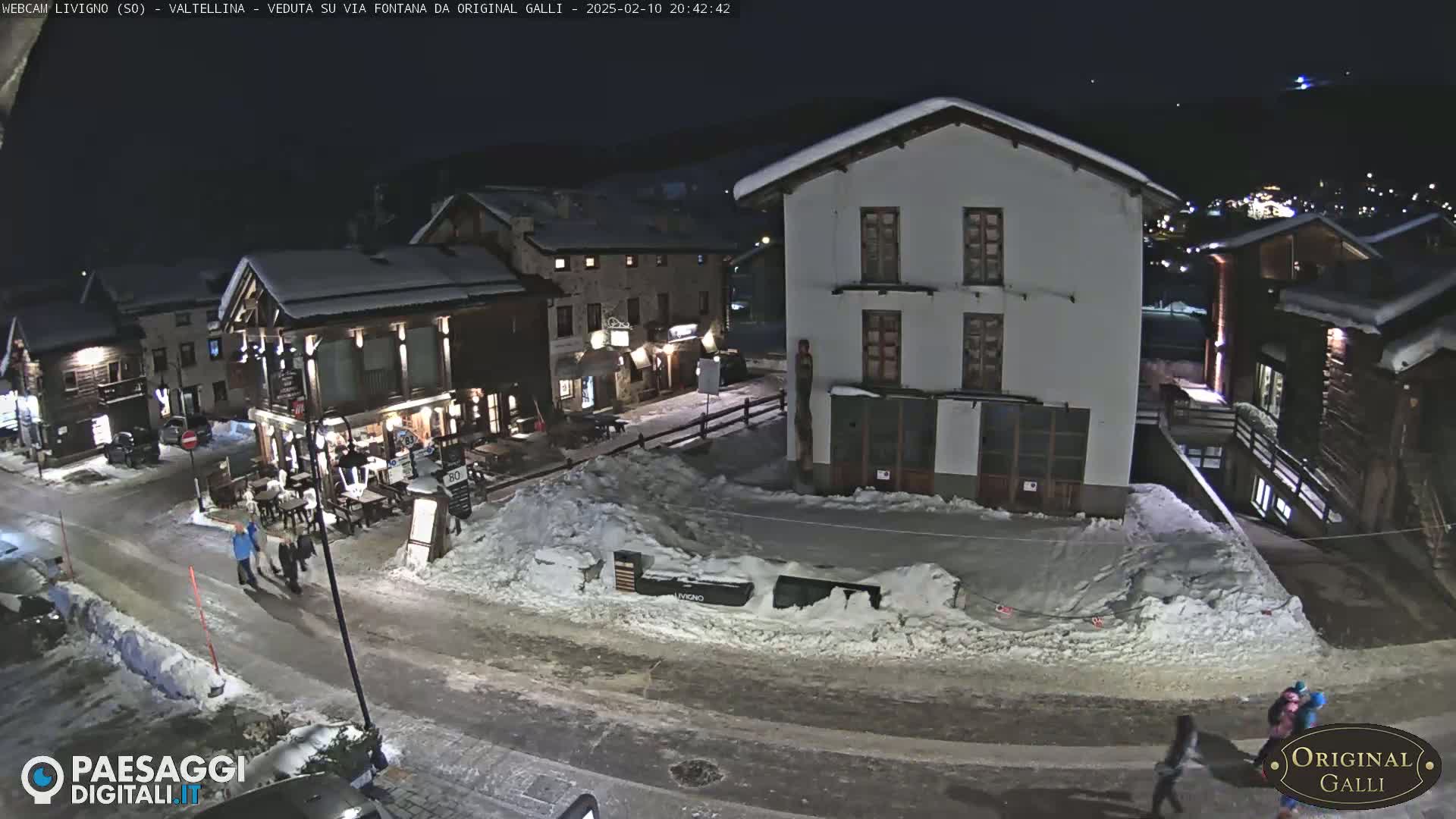 A snow-covered village street at night, with several buildings illuminated, and a few people walking.