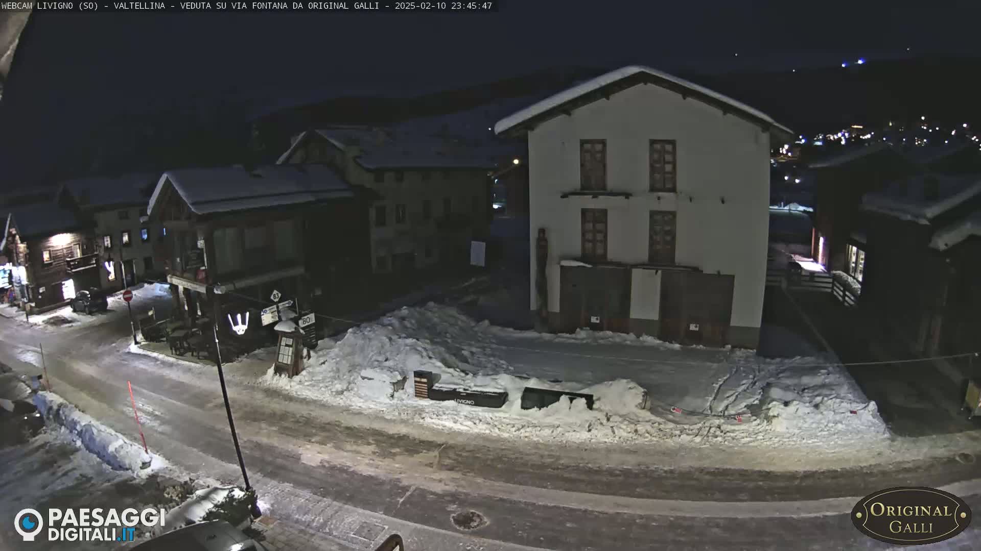 A snow-covered street at night in a village, with several buildings illuminated, under a dark sky.