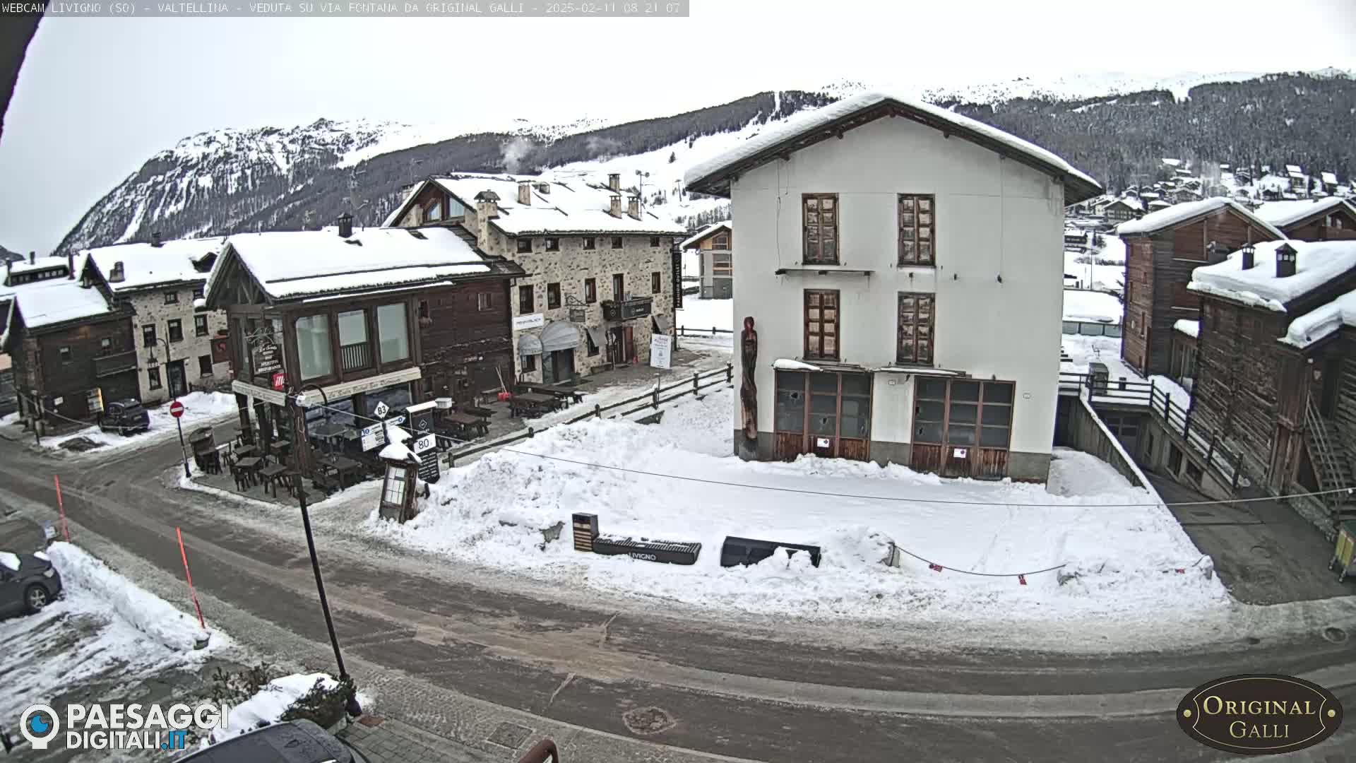 A snow-covered village square with several buildings, some with outdoor seating areas, is visible under an overcast sky.