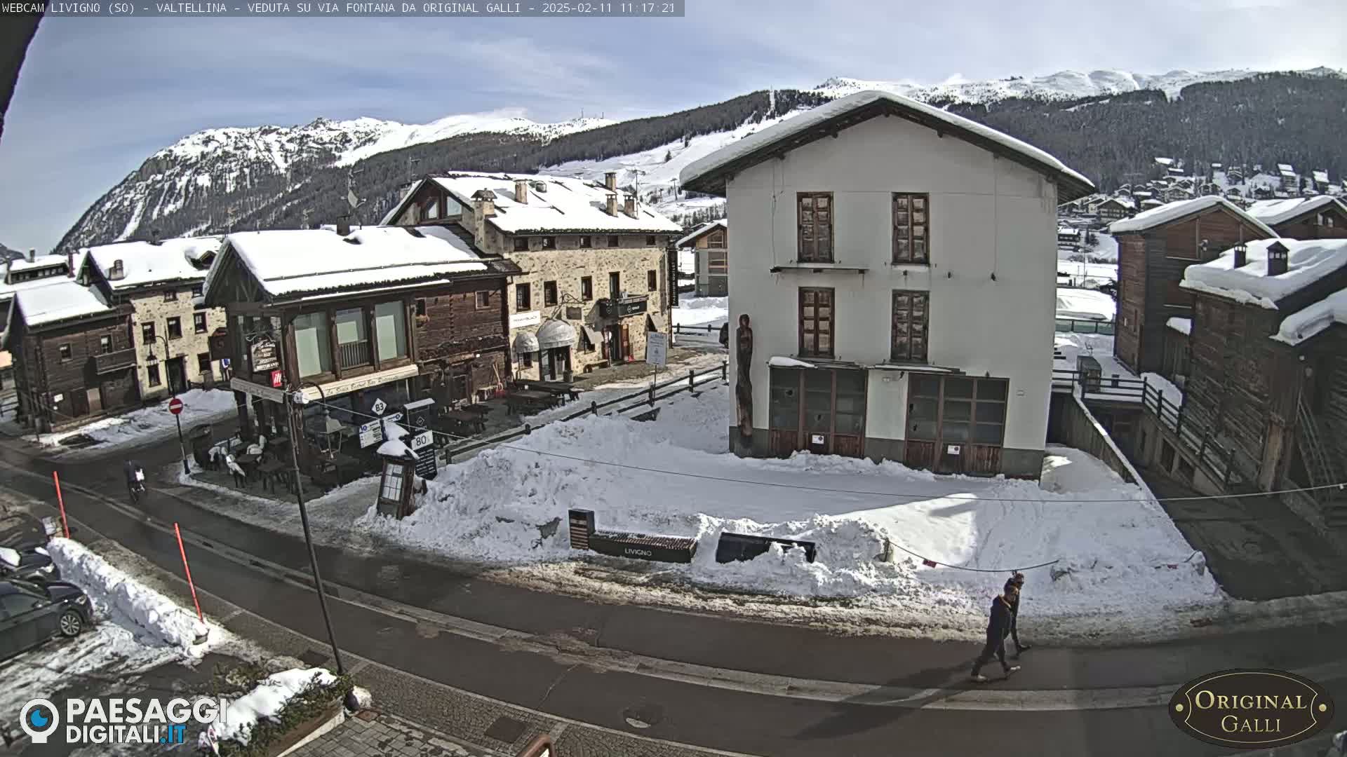 A snow-covered village street scene under a sunny sky, with several buildings, a person walking, and cars parked.