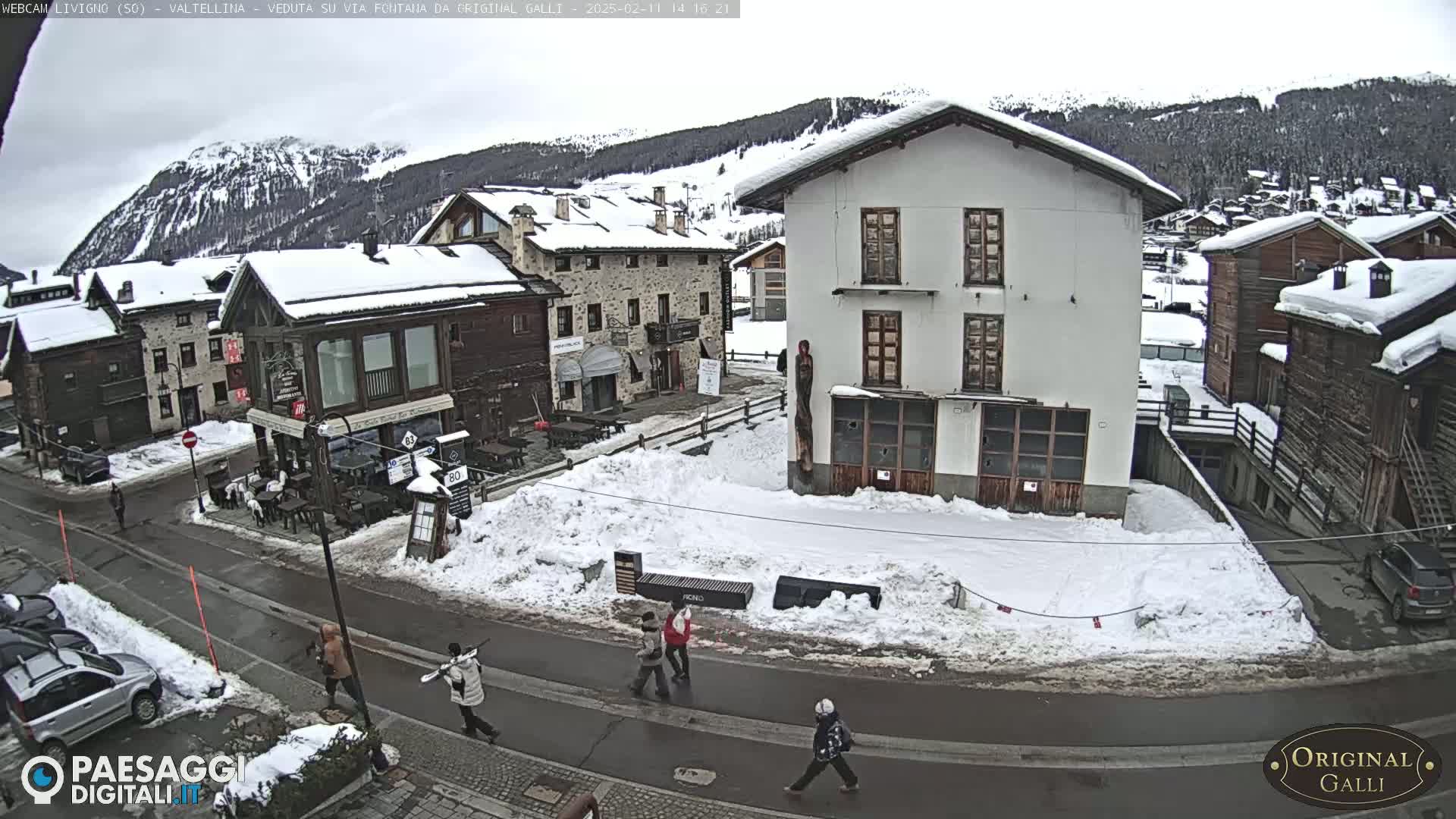 A snow-covered village street scene shows several people walking near buildings, with a mountain range in the background under an overcast sky.