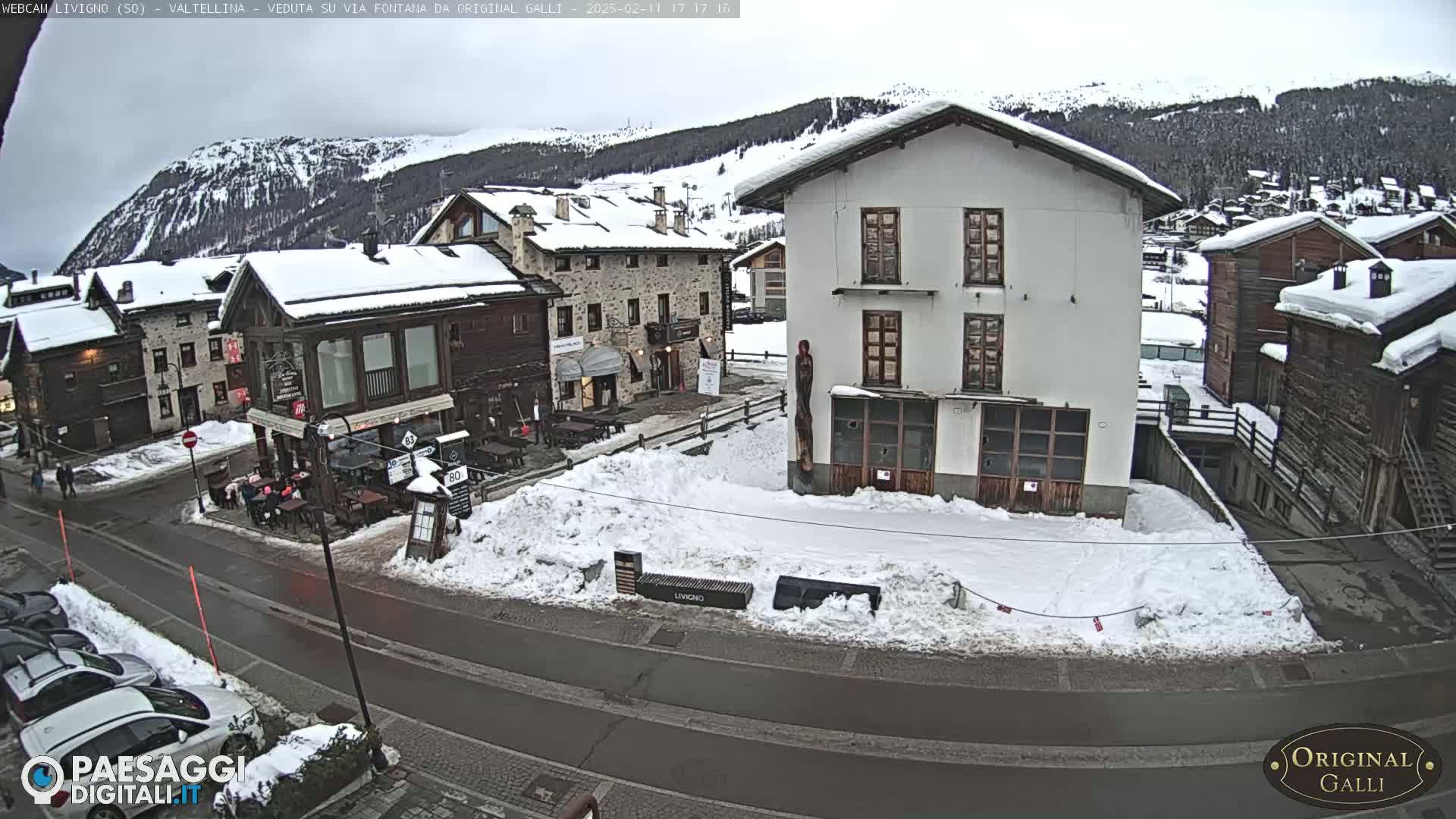 A snow-covered village scene shows several buildings, a street, and parked cars under an overcast sky, with snow-capped mountains in the background.