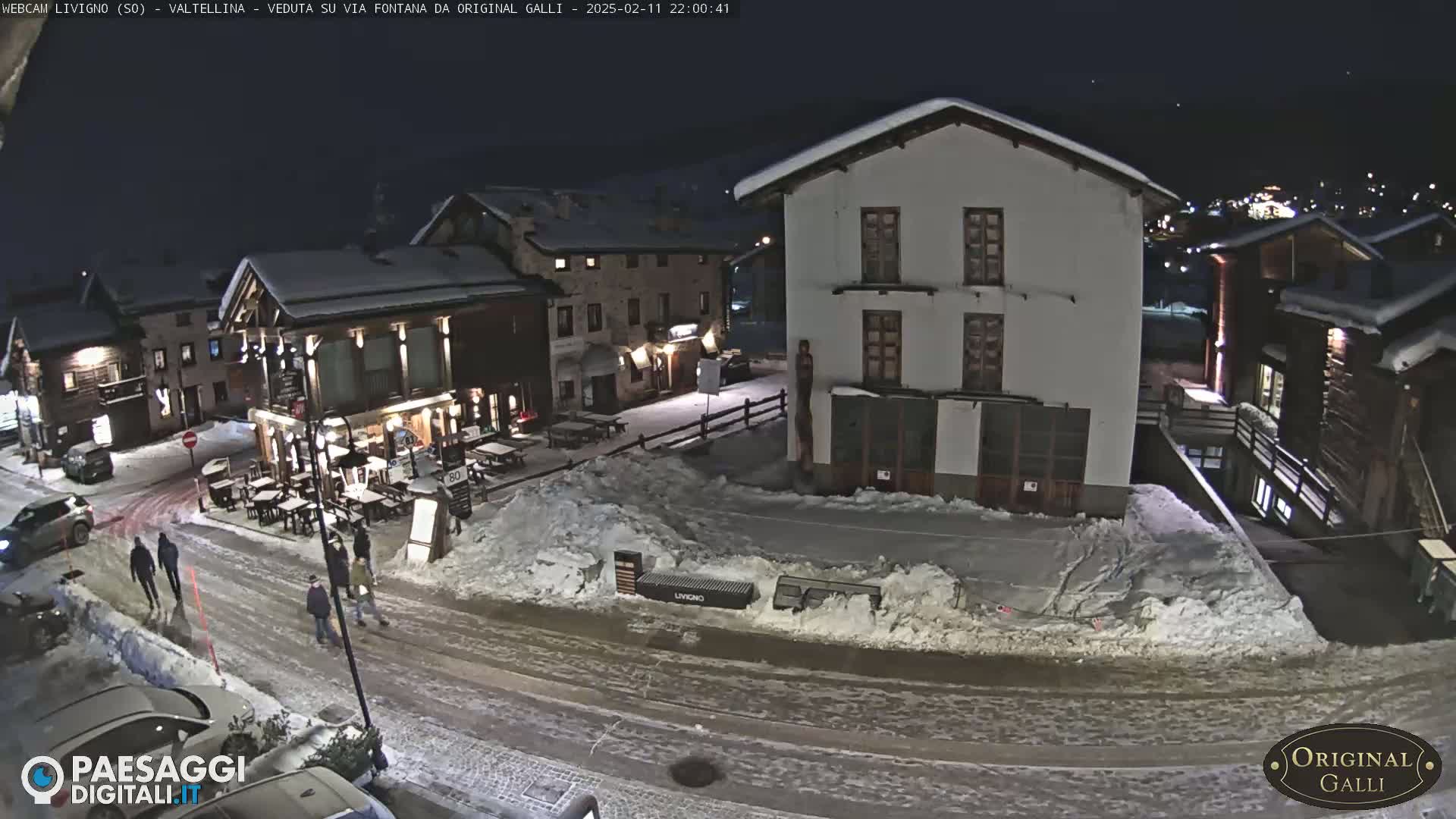 A snow-covered town square at night, with several buildings, a few people walking, and some cars parked.