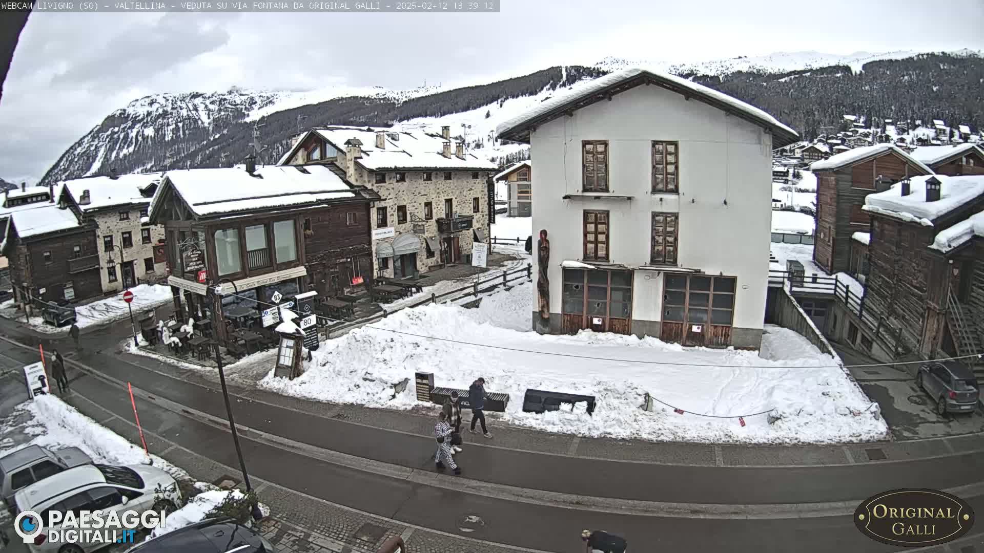 A snow-covered village street scene shows several buildings, some people walking, and parked cars under a cloudy sky.