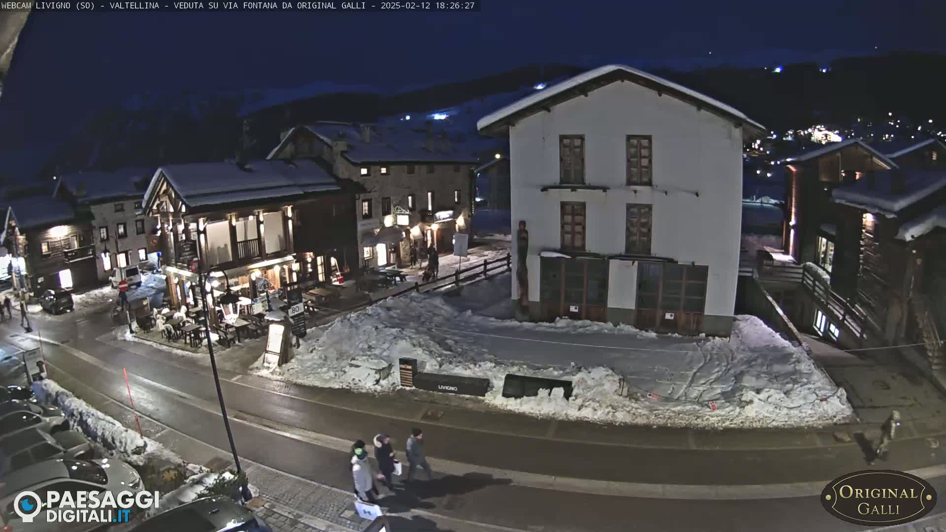 A snow-covered village street at night, with several buildings illuminated, and a few pedestrians walking.