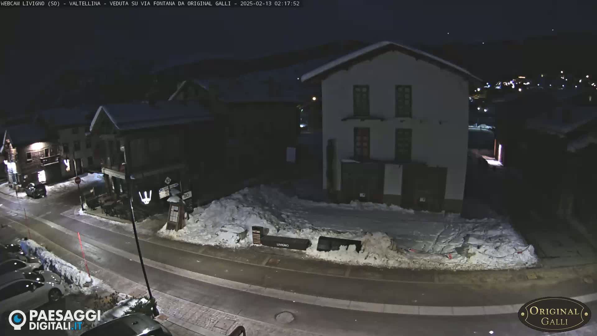 A snow-covered street at night in a village, with several buildings visible, including a two-story building and a restaurant with outdoor seating.