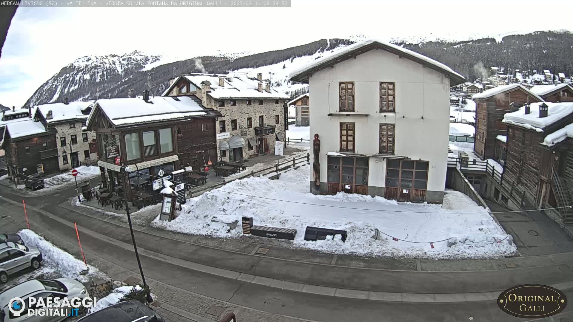 A snow-covered village street scene with several buildings, some with snow on their roofs, under an overcast sky.