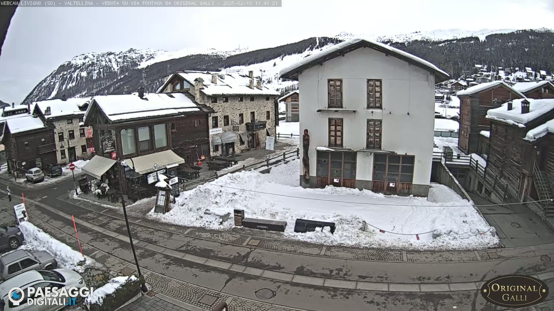 A snow-covered village square, nestled amongst snow-dusted buildings and mountains under an overcast sky.