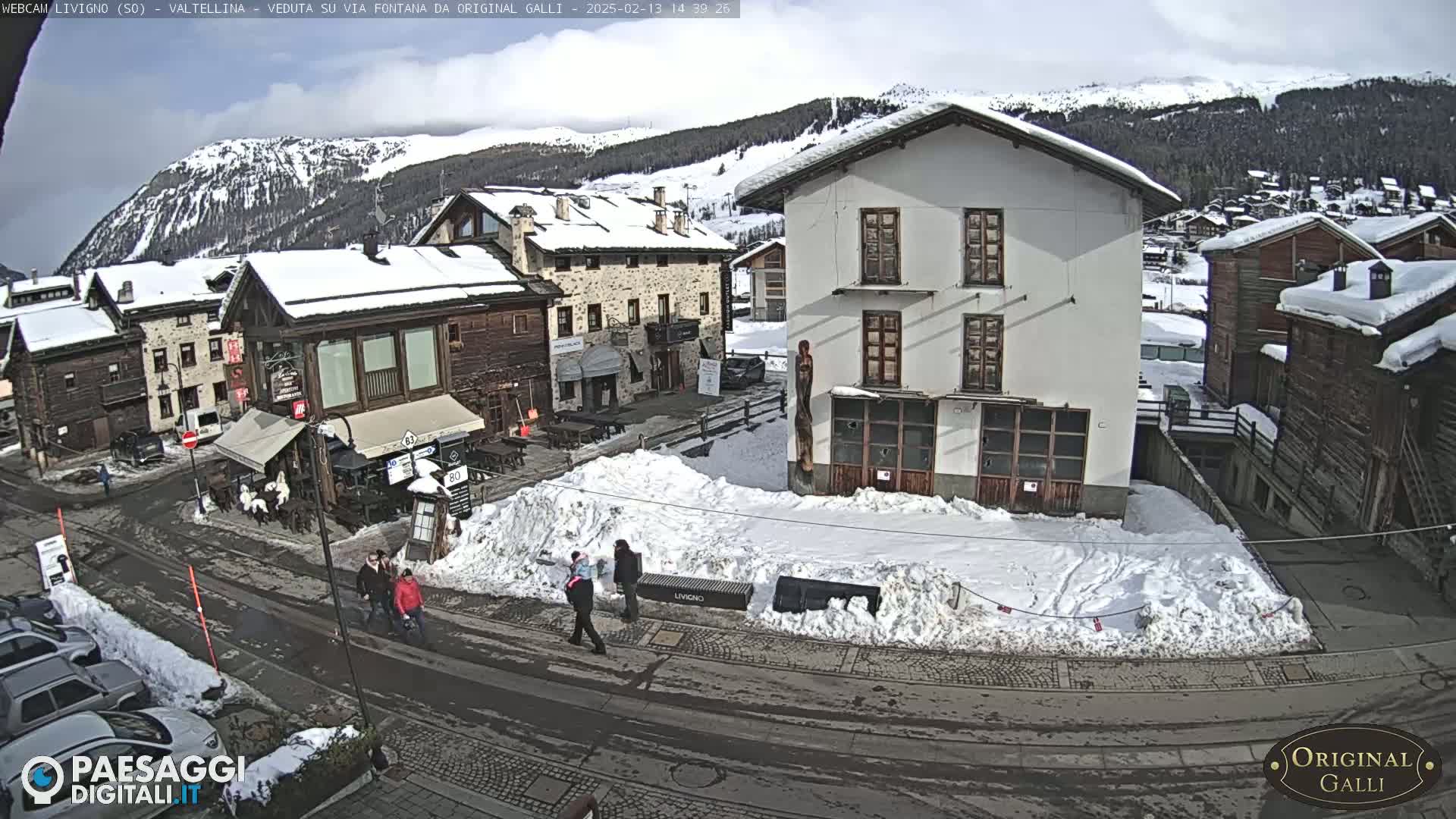 A snow-covered village street scene under a partly cloudy sky, with several pedestrians walking past buildings.