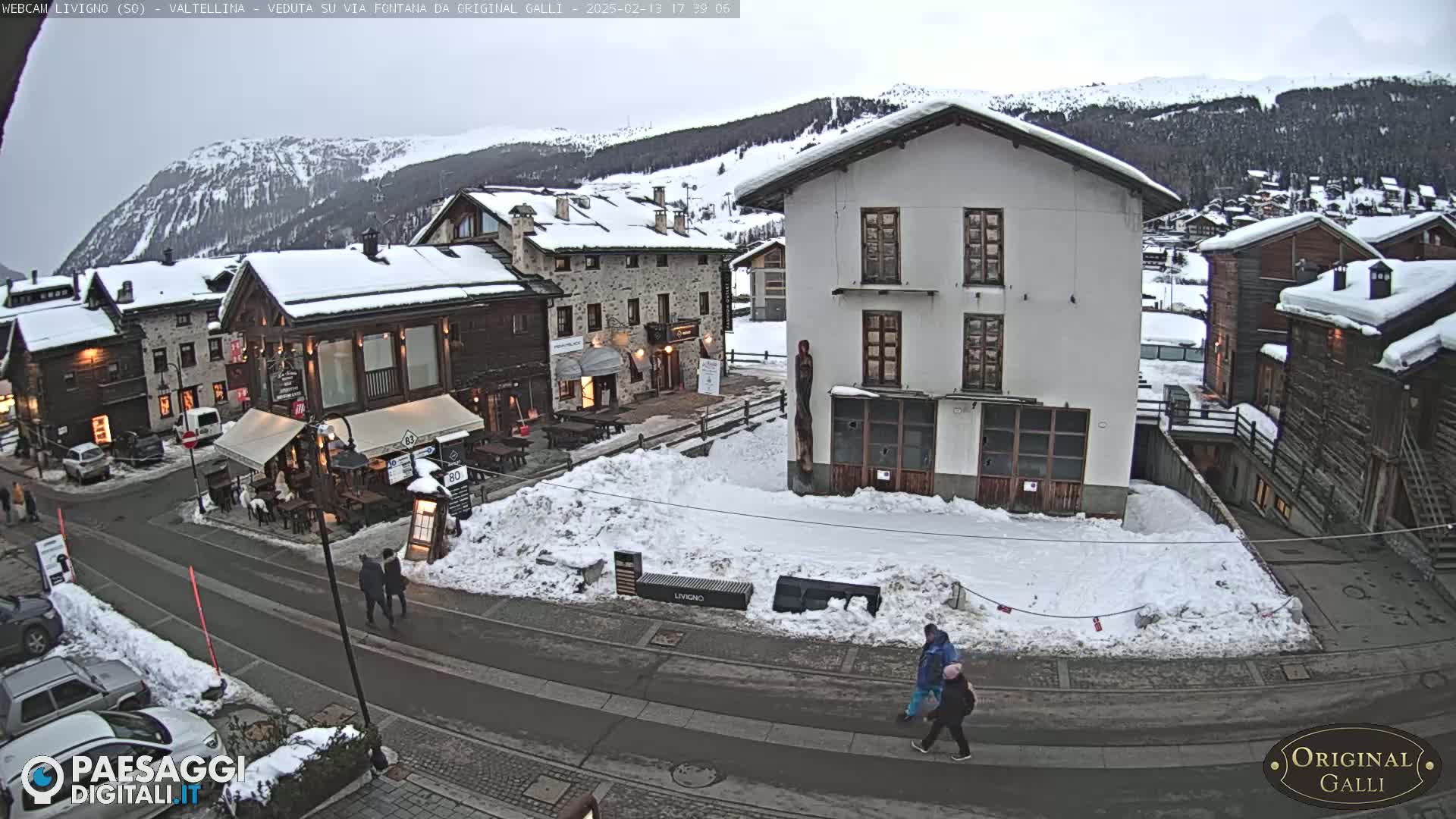A snow-covered village square with several buildings, some pedestrians, and cars parked on the street under an overcast sky.