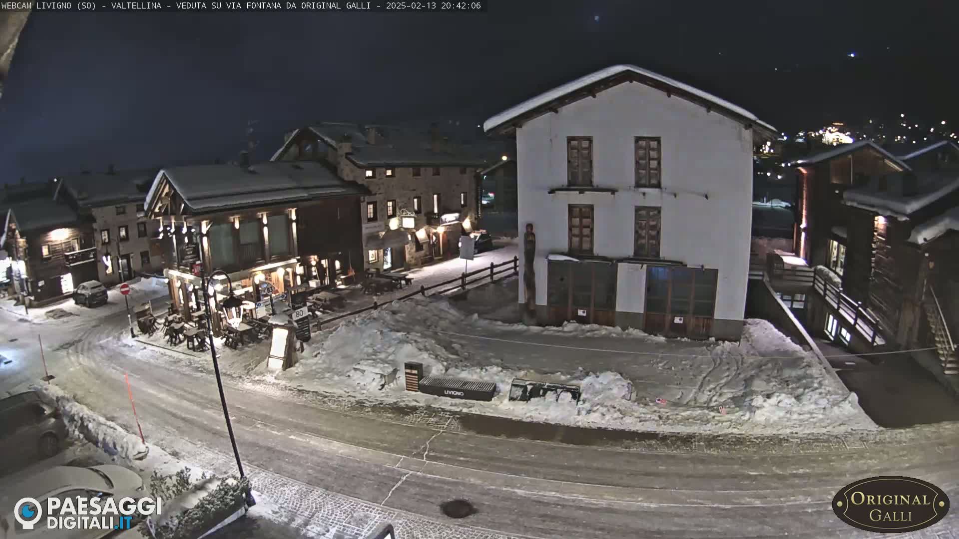 A snow-covered village street at night, with several buildings illuminated, shows a quiet scene under a clear, dark sky.