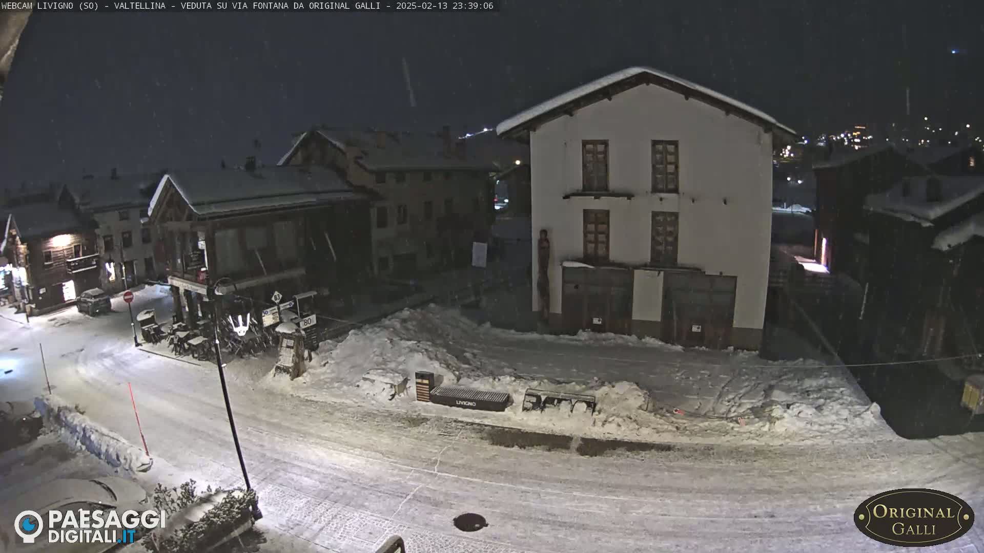 A snow-covered town square at night, lightly snowing, with several buildings visible.