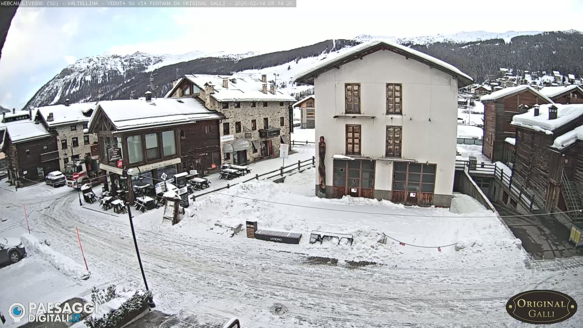 A snow-covered village square with several buildings, some with outdoor seating areas, is situated in front of a mountain range under a cloudy sky.