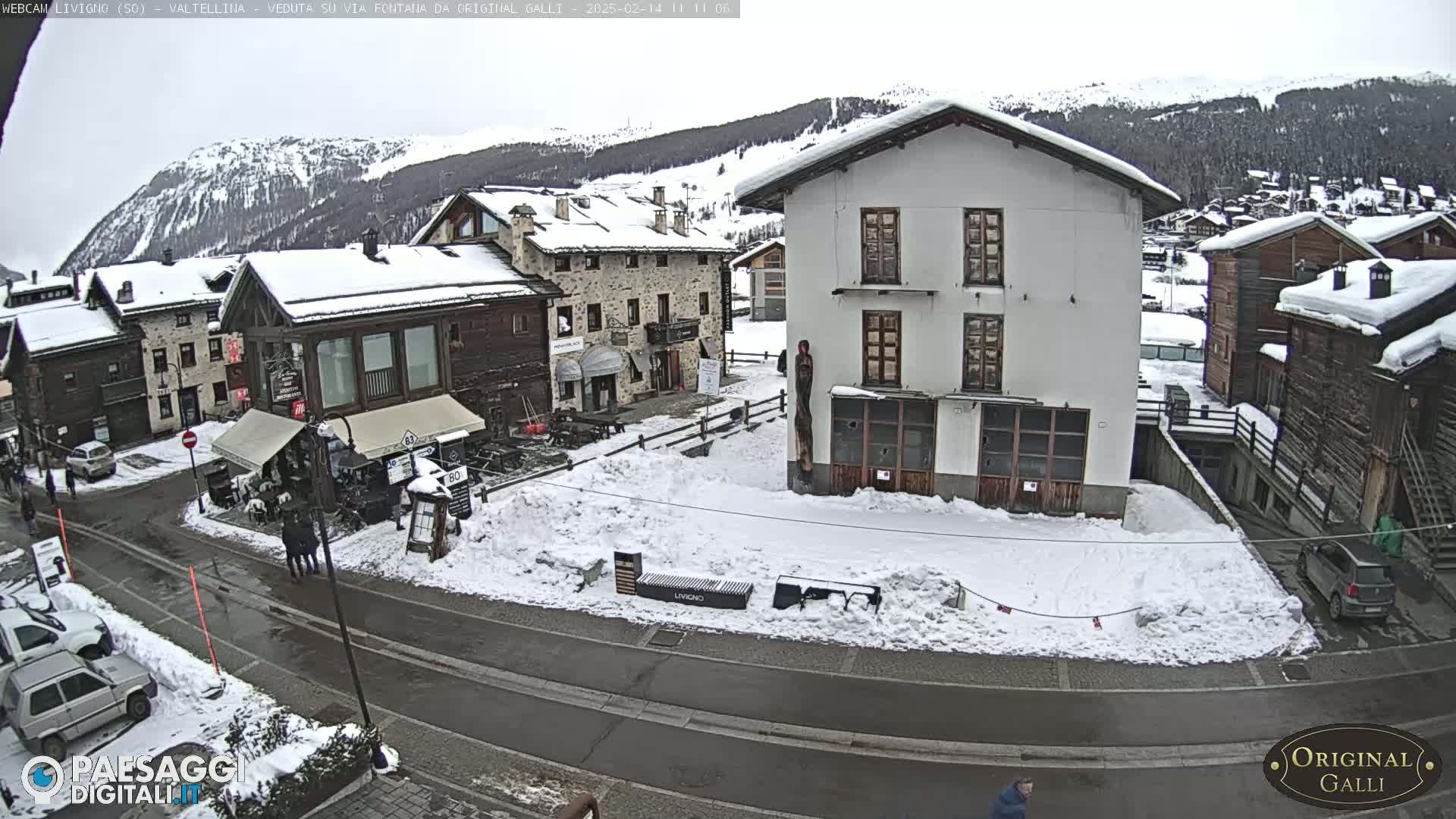 A snow-covered village square with several buildings, some cars parked, and a few pedestrians, under an overcast sky.