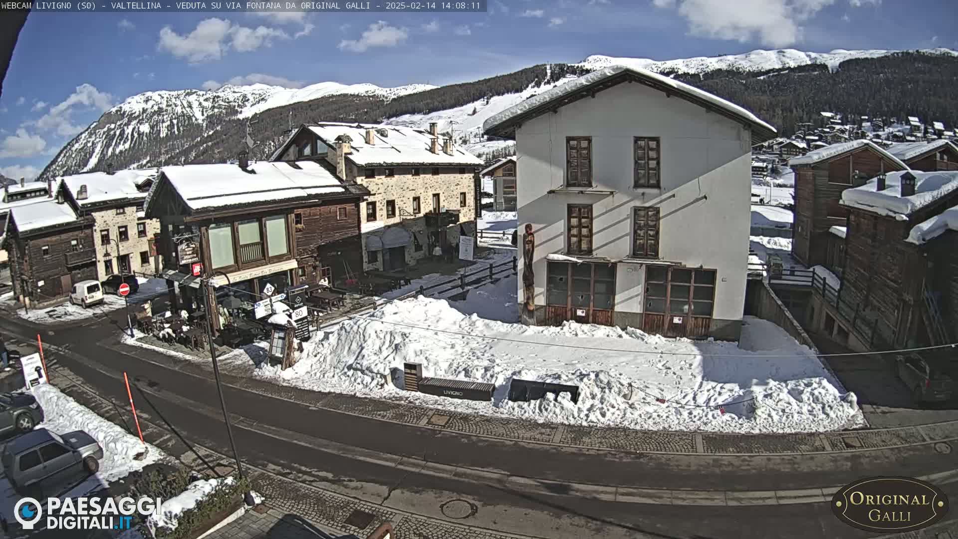 A snow-covered village square is viewed from above on a sunny day, with several buildings and parked cars visible.