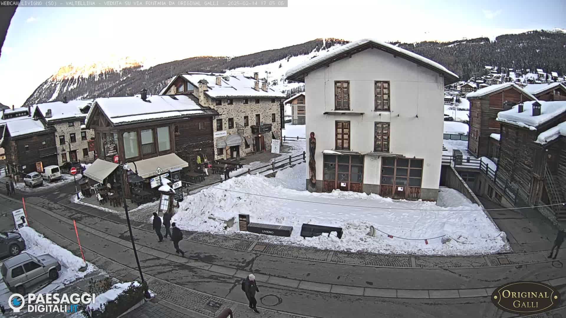 A snow-covered village street scene shows several stone buildings, some with snow on their roofs, and a few people walking on a road.