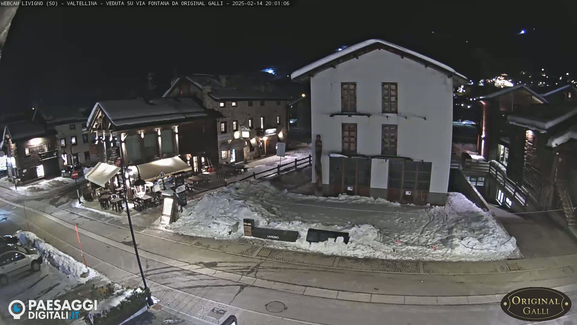 A snow-covered village square at night, with several buildings and a few cars parked nearby.
